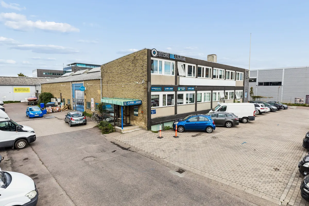 Flere biler parkeret på Bådehavnsgade i København. Kontorbyggeri med skrift Byens Billeje.dk. Solrig himmel med skyer. Stiger med højt fremspring.