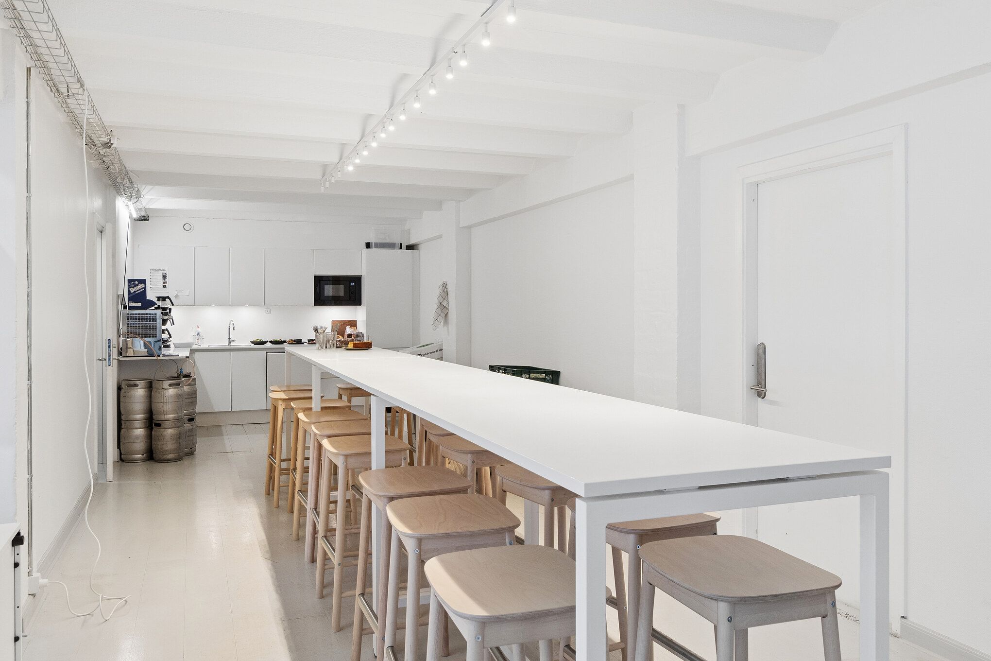 Minimalistisk kontor med hvidt skrivebord og stole og lyse loftsbjælker. Der er højt gulv og store vinduer med dagslys. Der er køleskab og mikroovn bagved.