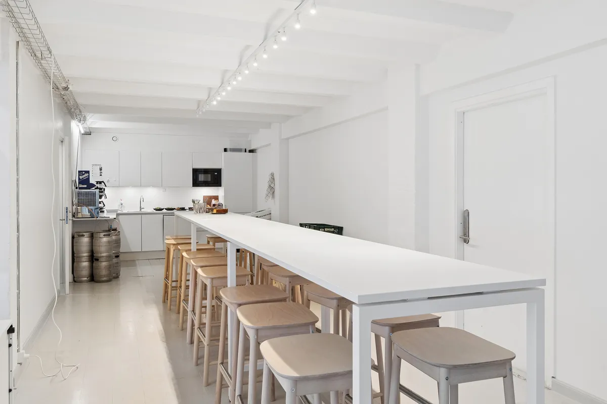 Minimalistisk kontor med hvidt skrivebord og stole og lyse loftsbjælker. Der er højt gulv og store vinduer med dagslys. Der er køleskab og mikroovn bagved.