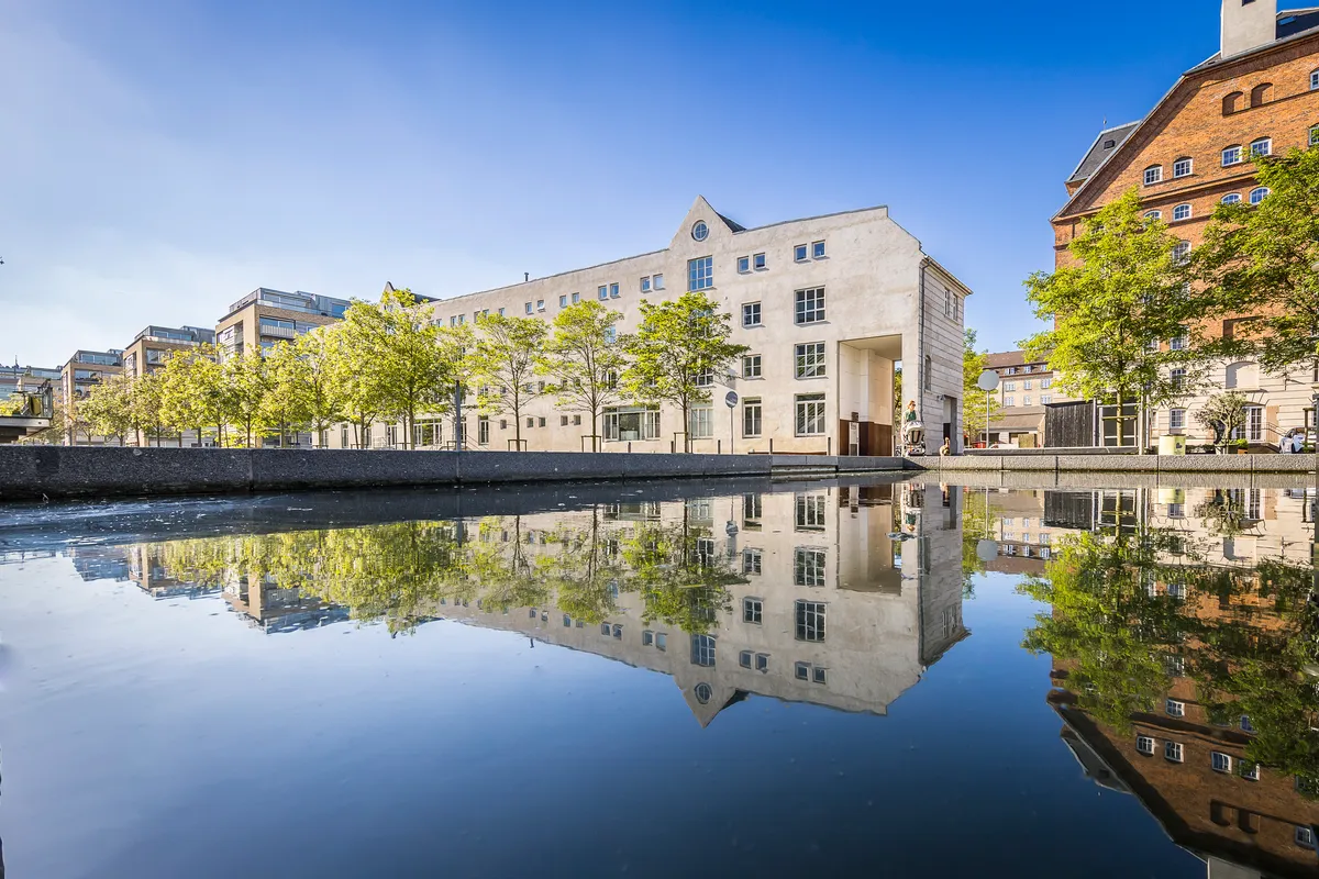 Lyst kontormiljø med masser af dagslys, lyse murstensvægge og store glasvinduer. Refleksioner af bygningen og træer i vandet