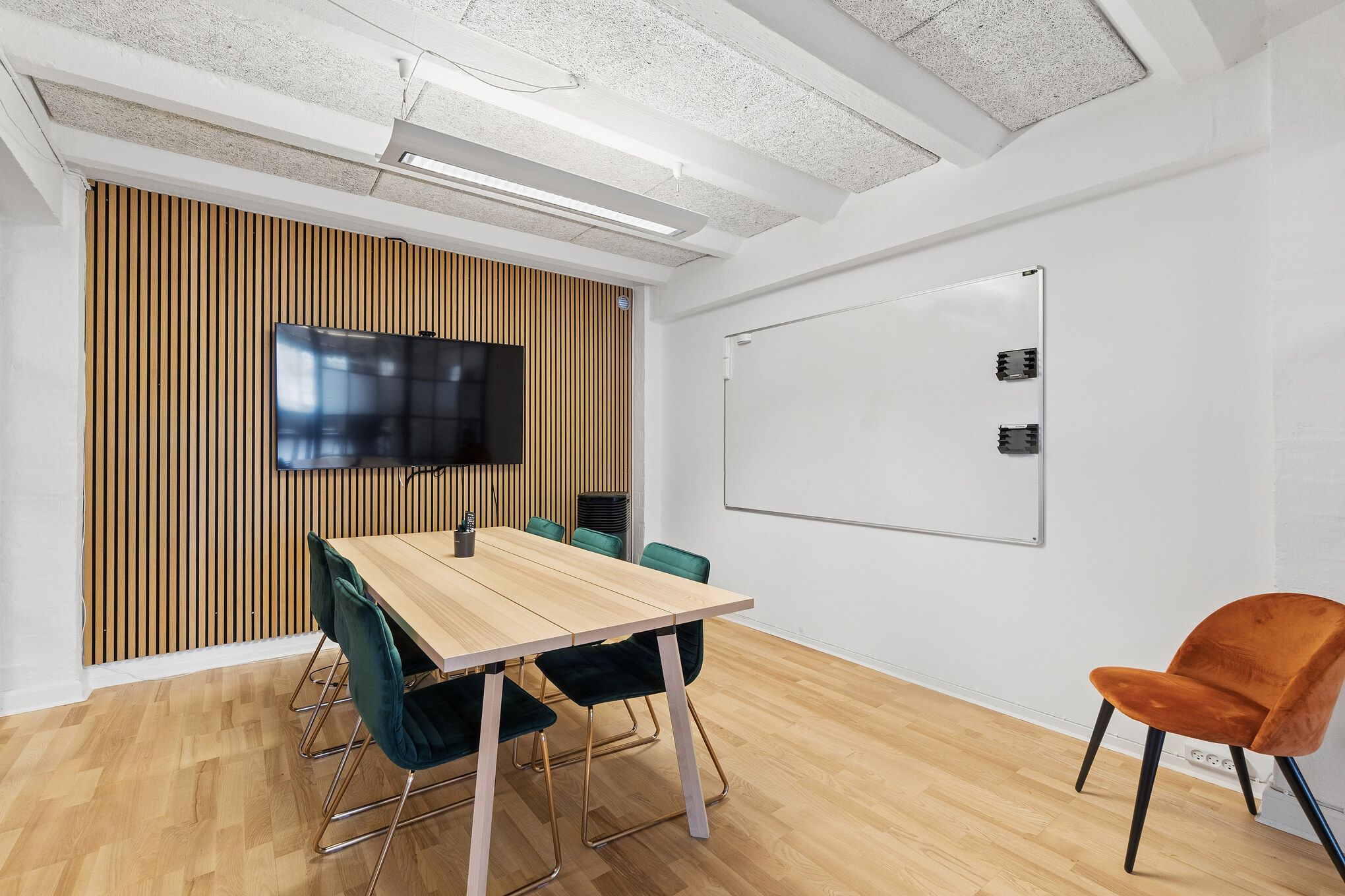 Lyst og indbydende mødelokale med åben gulvplan og træbord. Højt loft med hvidt og træelementer samt lyse hvide vægge. Bord med moderne grønne stole og hvid tavle.