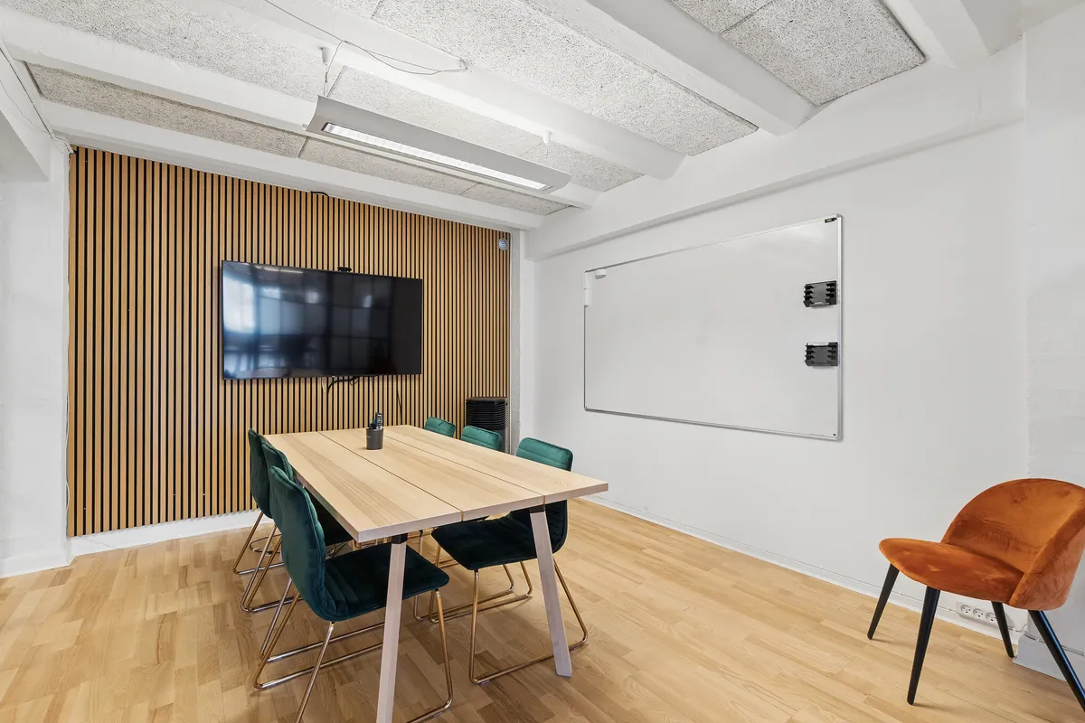 Lyst og indbydende mødelokale med åben gulvplan og træbord. Højt loft med hvidt og træelementer samt lyse hvide vægge. Bord med moderne grønne stole og hvid tavle.