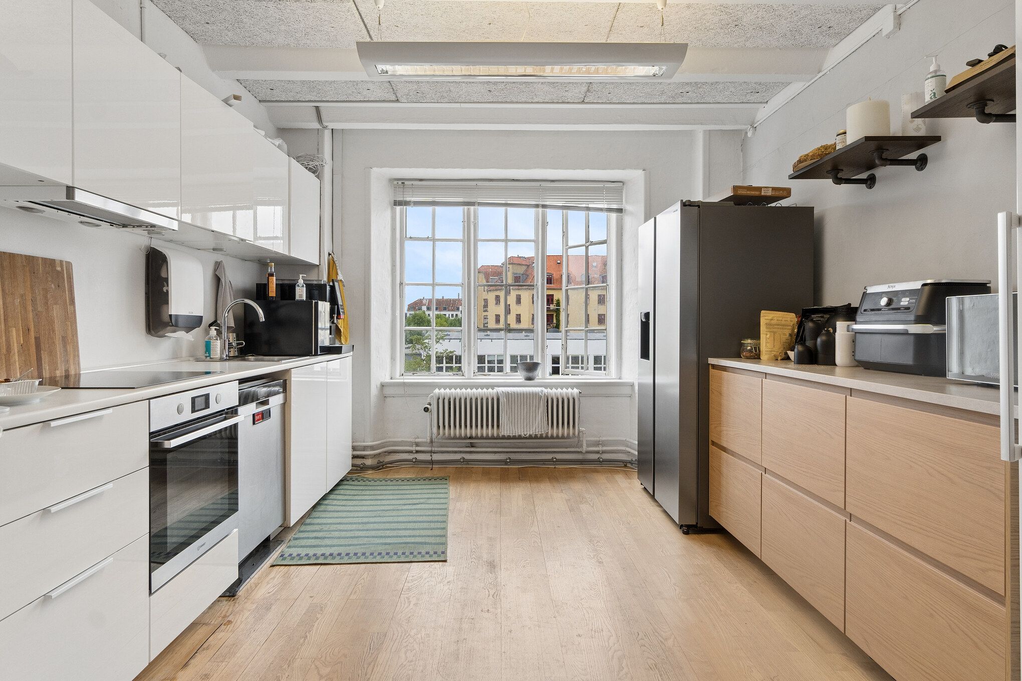 Et lyst og indbydende kontor med lyse kontorrummer, hvide køkkenskabe, moderne apparater og en terrasse med udsigt til byen, alle udstyret med lyse vinkler og lyse vinduer