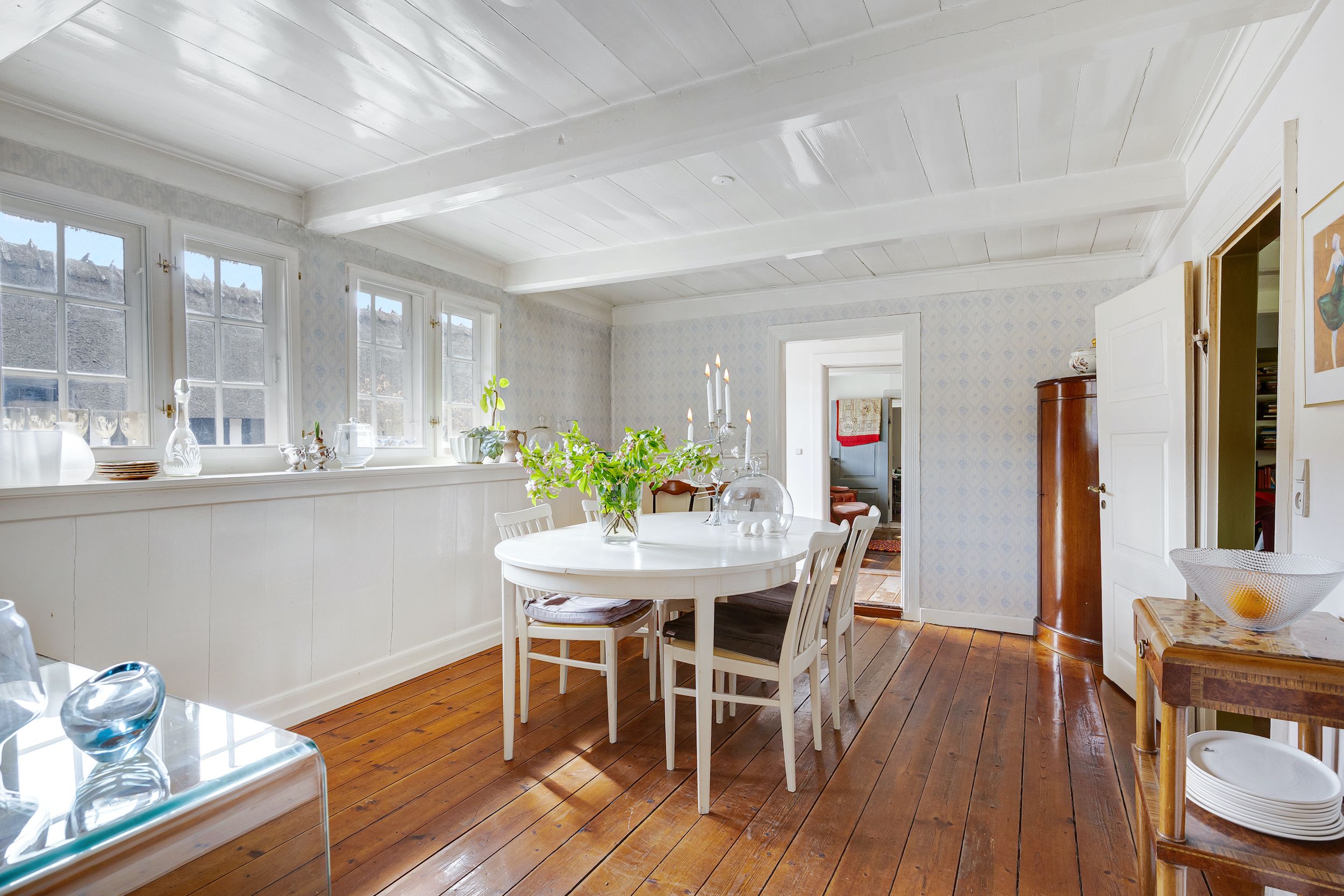 En moderne, hvid malet spisesal med trægulve og hvide stole omkring et rundt bord med blomster og et lyseskind