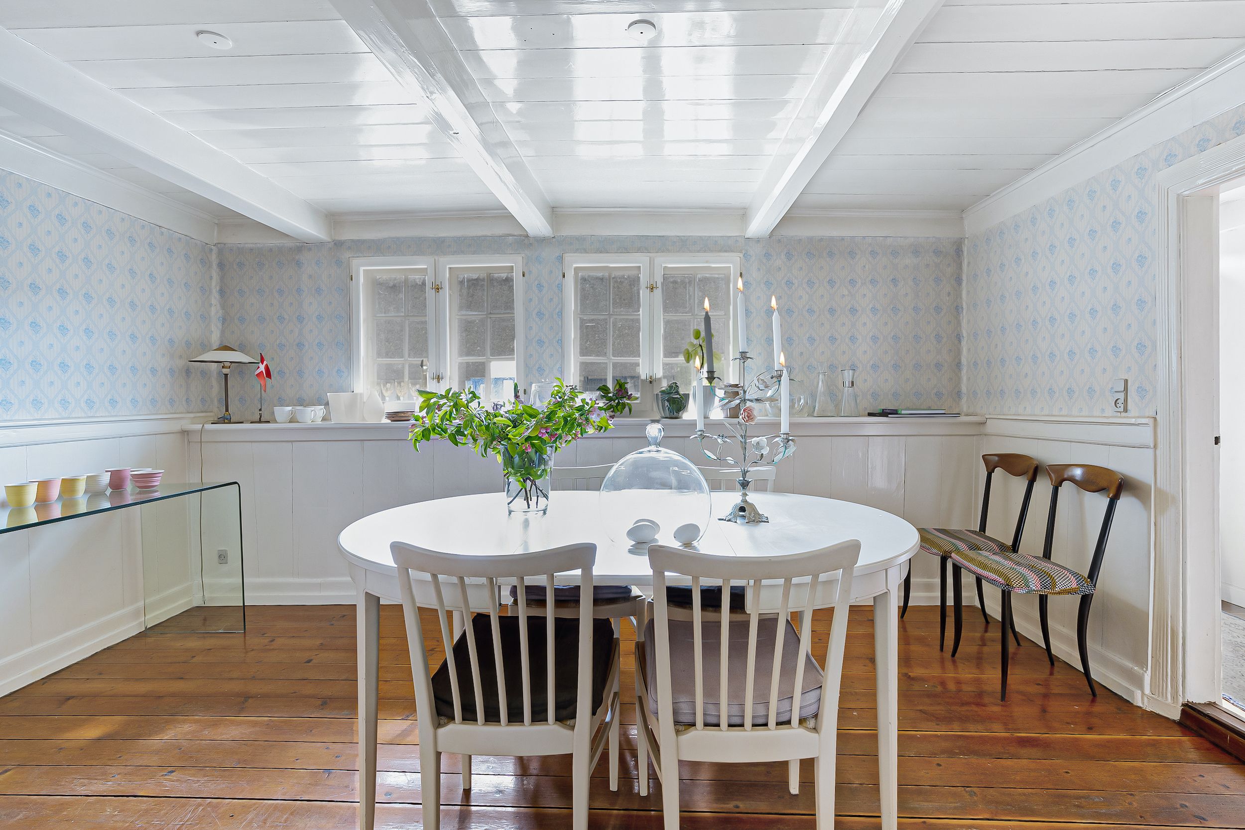 En hyggelig spisestuee med hvidt bord og stole, hvid loftsbeklædning og møbler, lysekroner og et glasbord med krus og