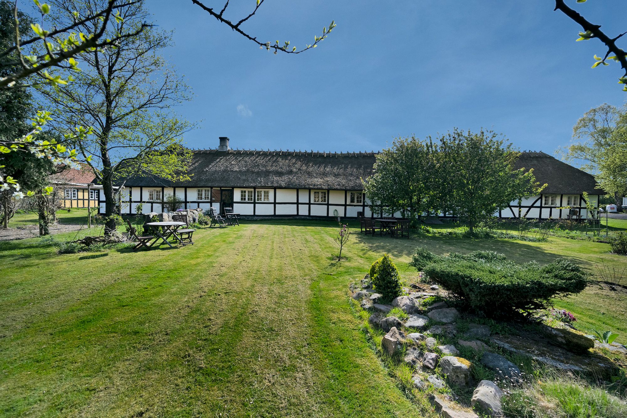 Gård med nyttehave, træer og en have med lave buske, placeret på en stor grøn eng med en hyggeligt indrettet hus og en skov