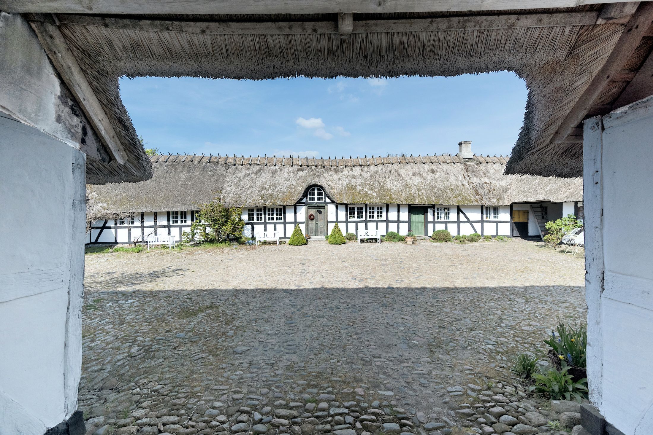 Fredet landsted med nyttehave og træklæsset tag og hvide vægge. En række hvide bænke står foran bygningen.