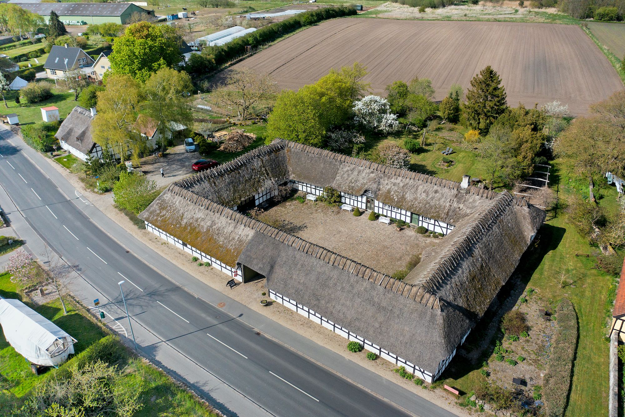 Feriebolig med tagdækket af halm ved siden af en asfaltvej og med grønne marker og skove i baggrunden