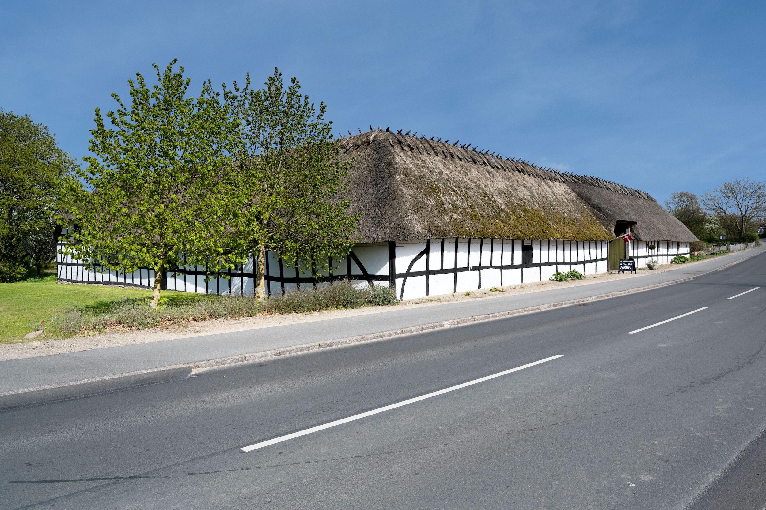 En gammel, hvid og sort malet gård med en stor, grøn, mosbevokset stråtag. En lille autoværkstedsskilt og en flagstav