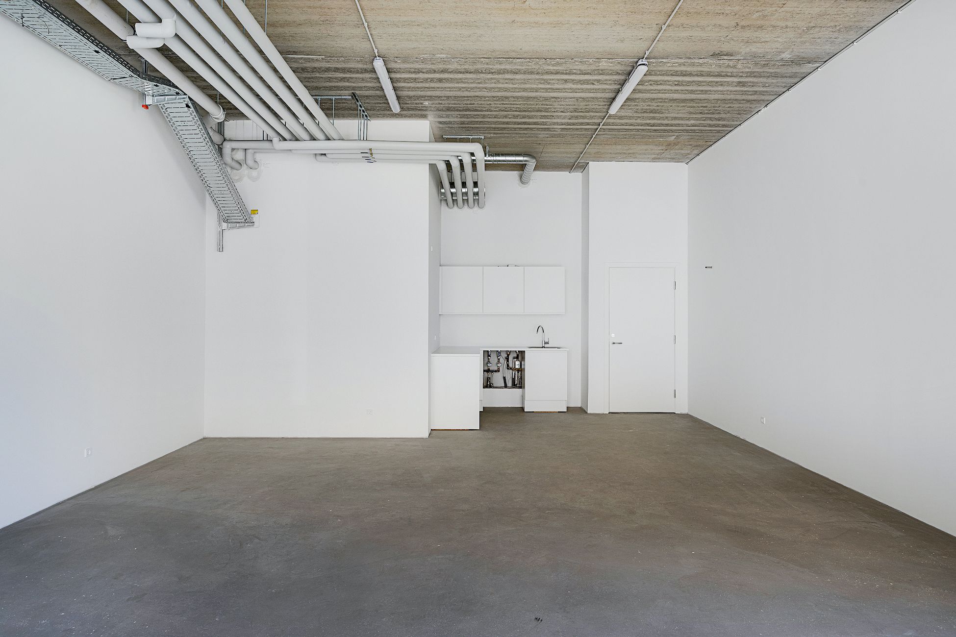 En tom rum med lyse vægge og cementgulv og et lille køkkenhjørne med håndvask og apparater bag en hvid skur