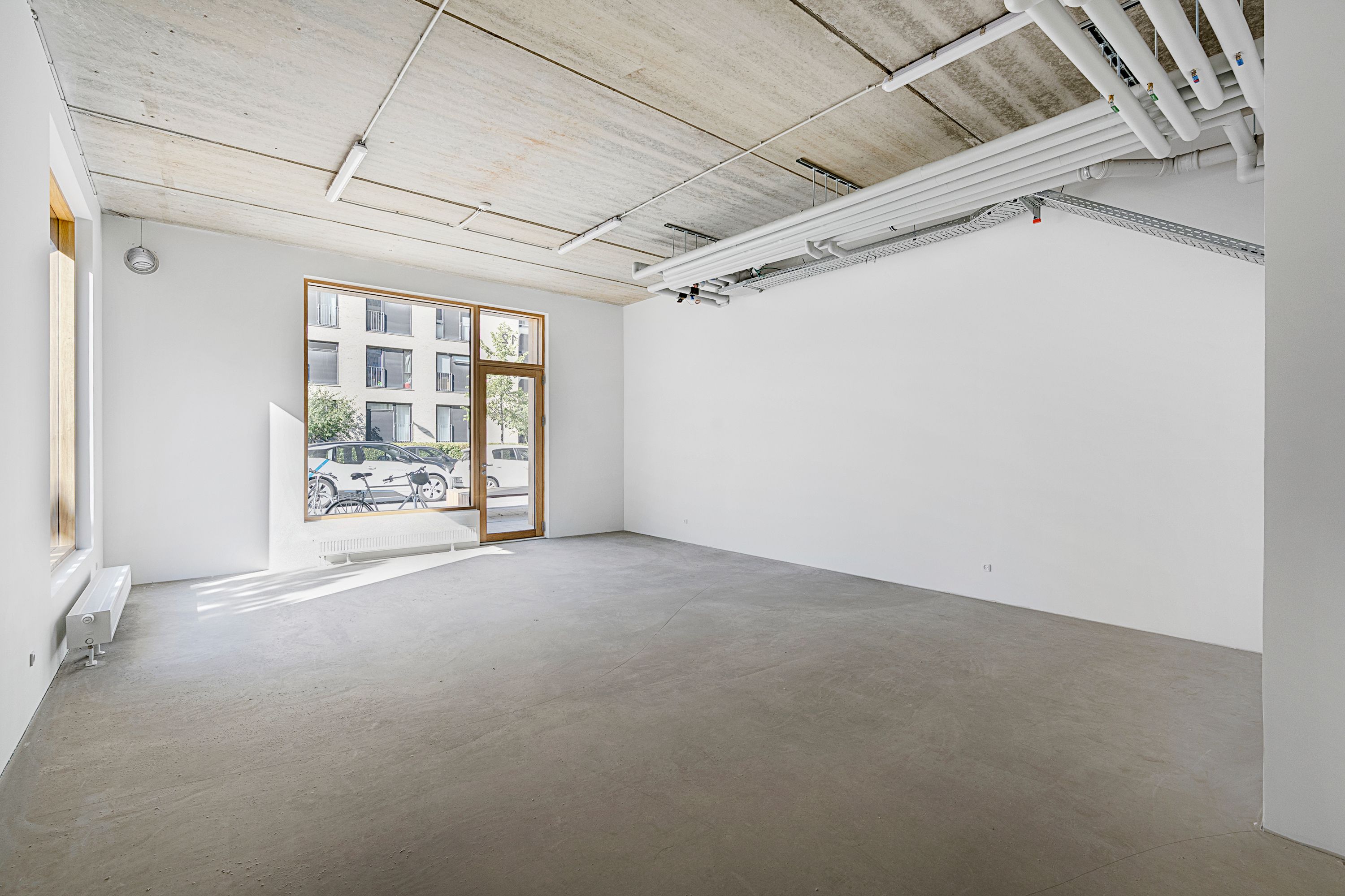 Rummelig lejlighed med vinduer mod gaden og solskin uden for. Betongulv og hvide vægge samt lysekroner på loftet