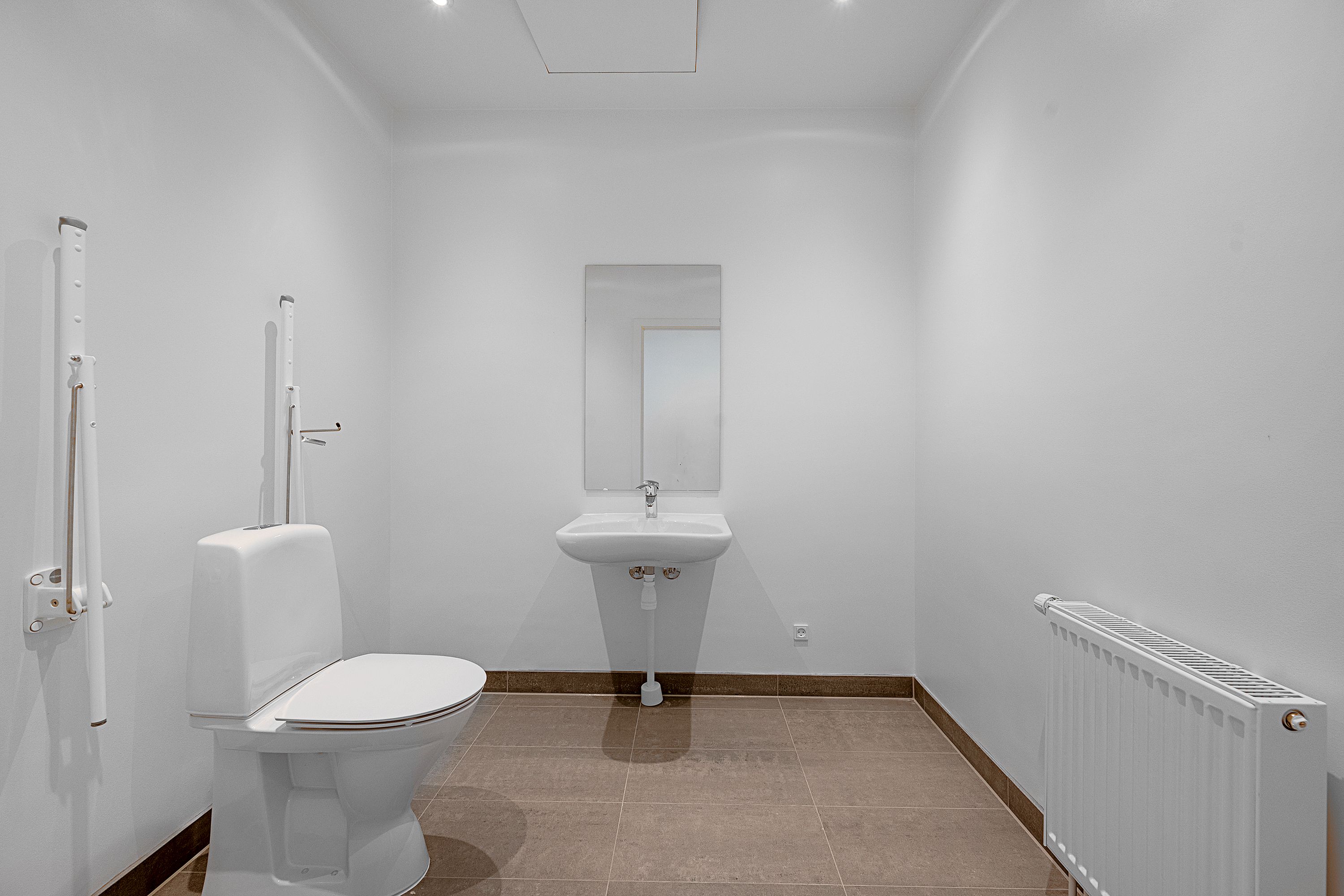 Et lille, hvidt badeværelse med en toilet, et hængende spejl og en radiator. Gulvet er dækket med lysegrå fliser og væggene