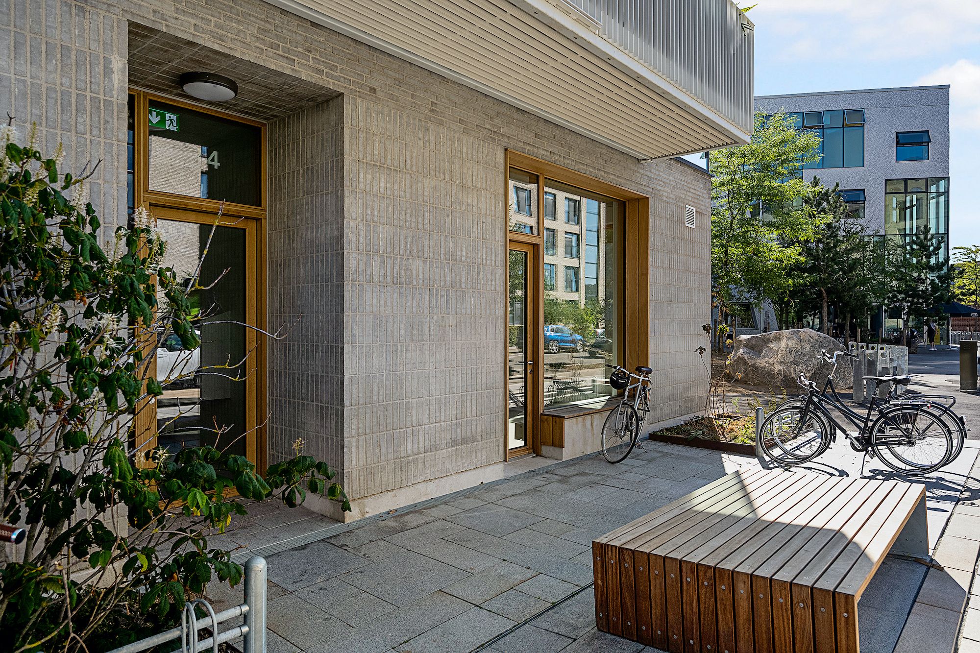 Lejlighed med stor indgangsdør og vindue, to parkerede cykler, skovle, plante og bænk udenfor bygningen