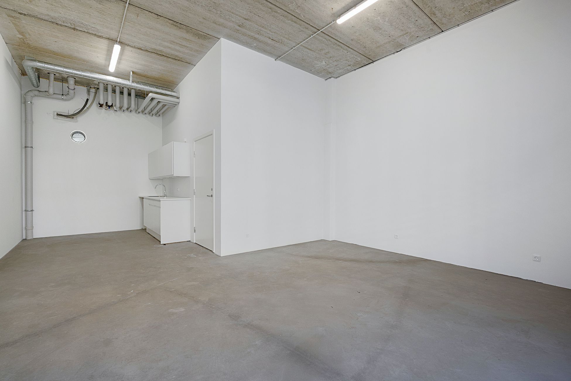 Et tomt rum med lyse hvide vægge, et køkken med lysegrå skabe og en hvid køkkenblok samt en hængende pibe på væggen