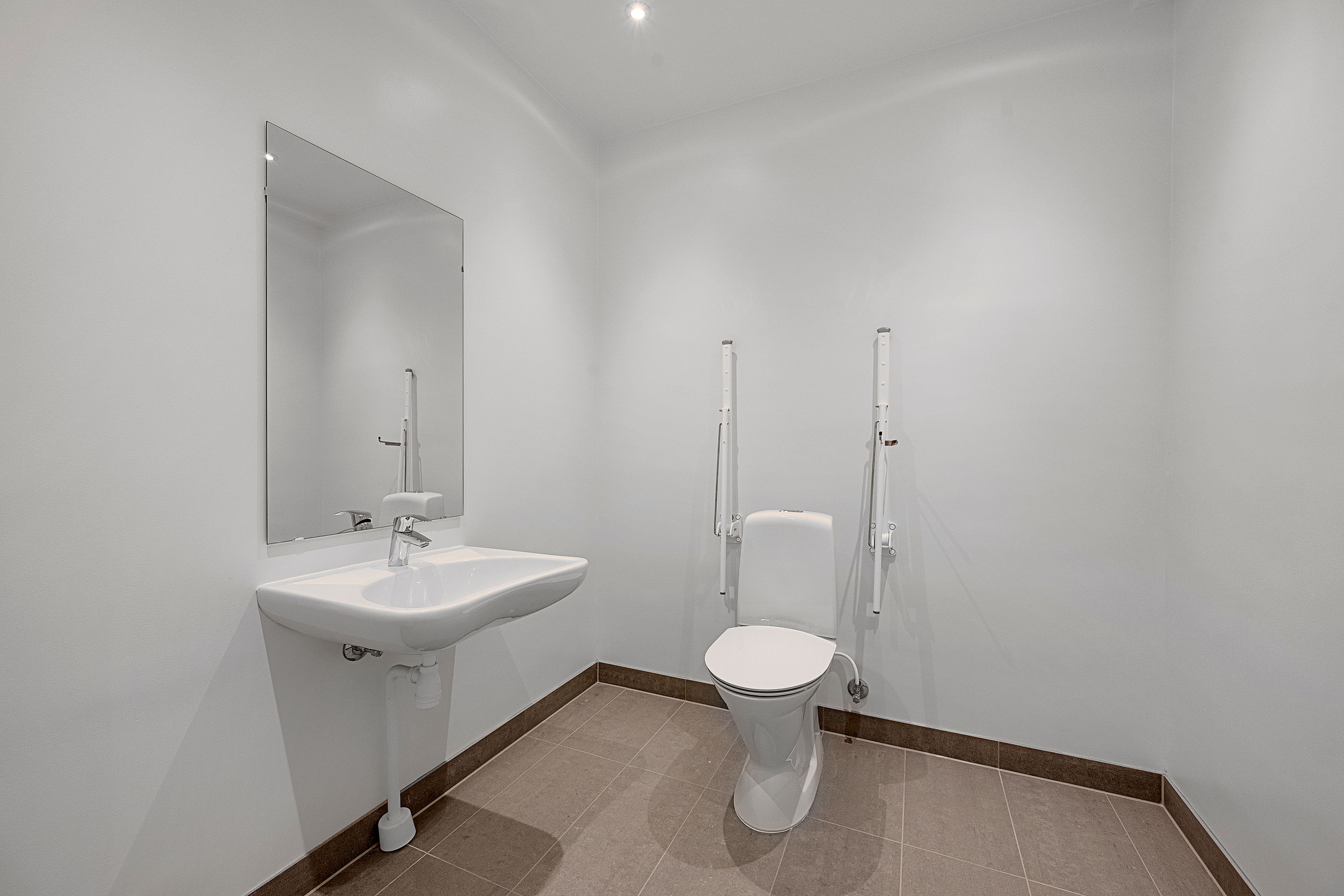 Et hvidt, funktionelt badeværelse med en hvid toilet, en hvid sink og en spejl, der reflekterer den hvide væg og loft