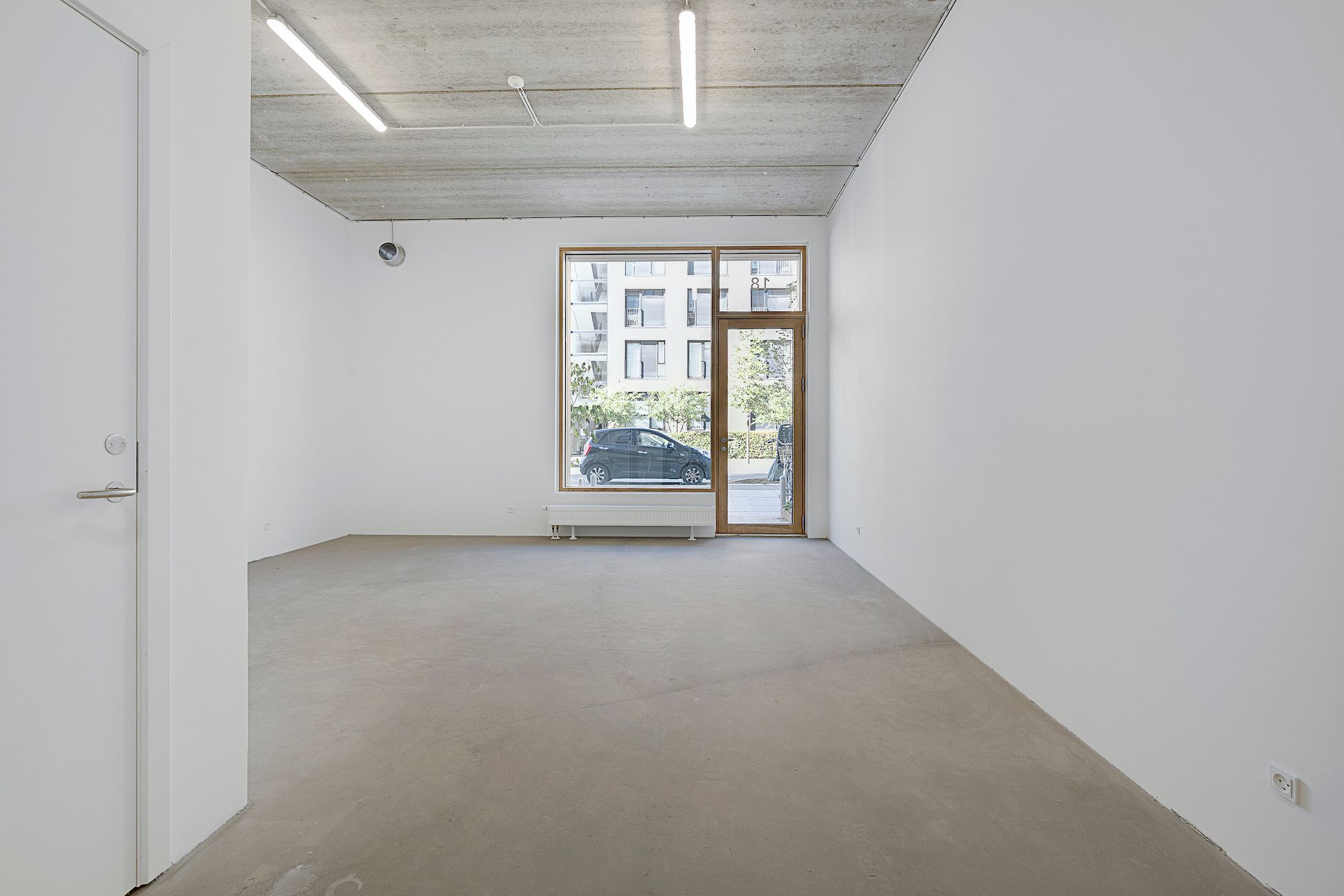 En stor, tom lejlighed med hvide vægge og cementgulv samt et stort vindue og et dørkar