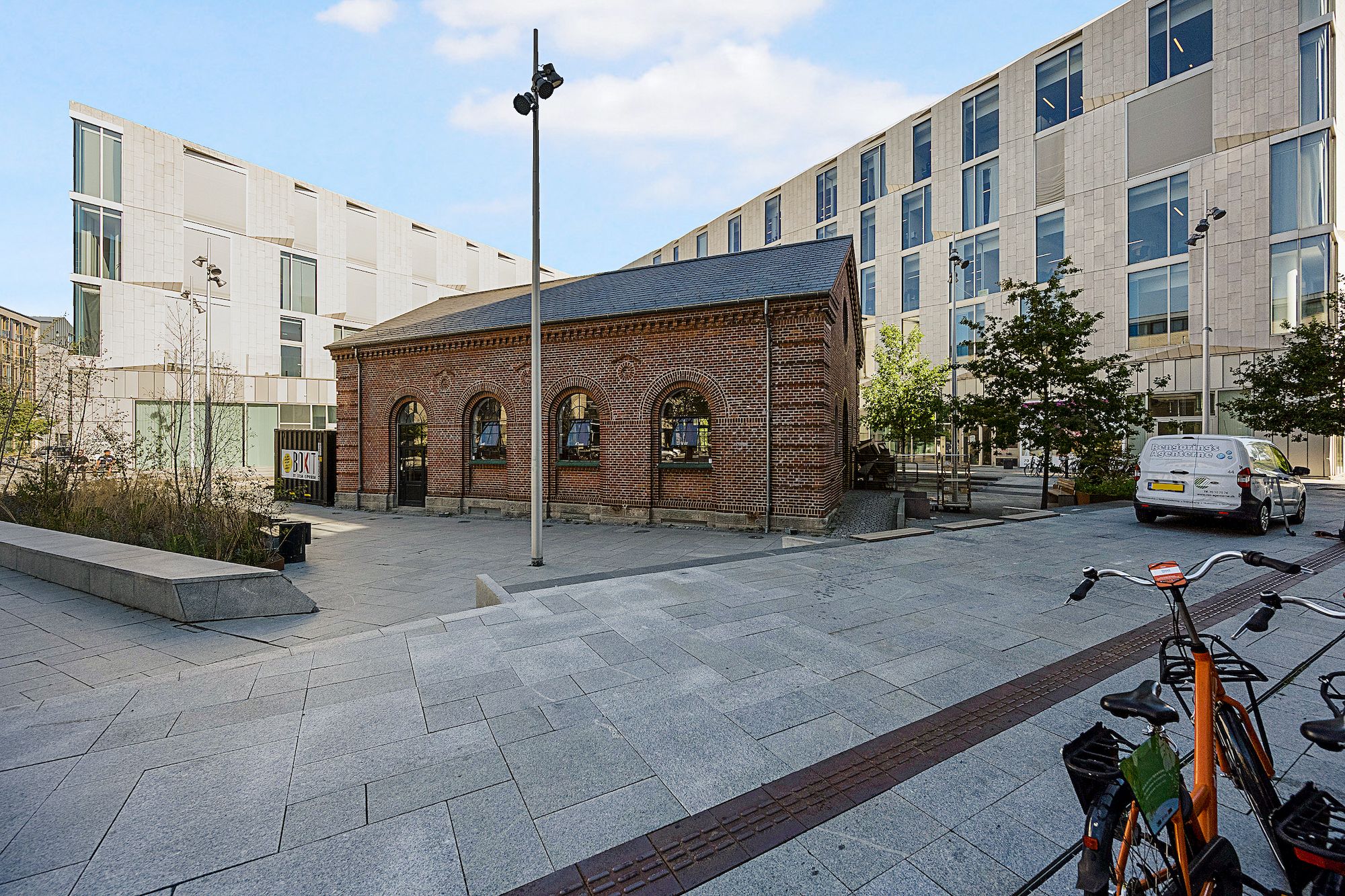 Lejlighedsbyggeri med røde mursten og vinduer i en bypark med grønne træer og skulpturer i baggrunden