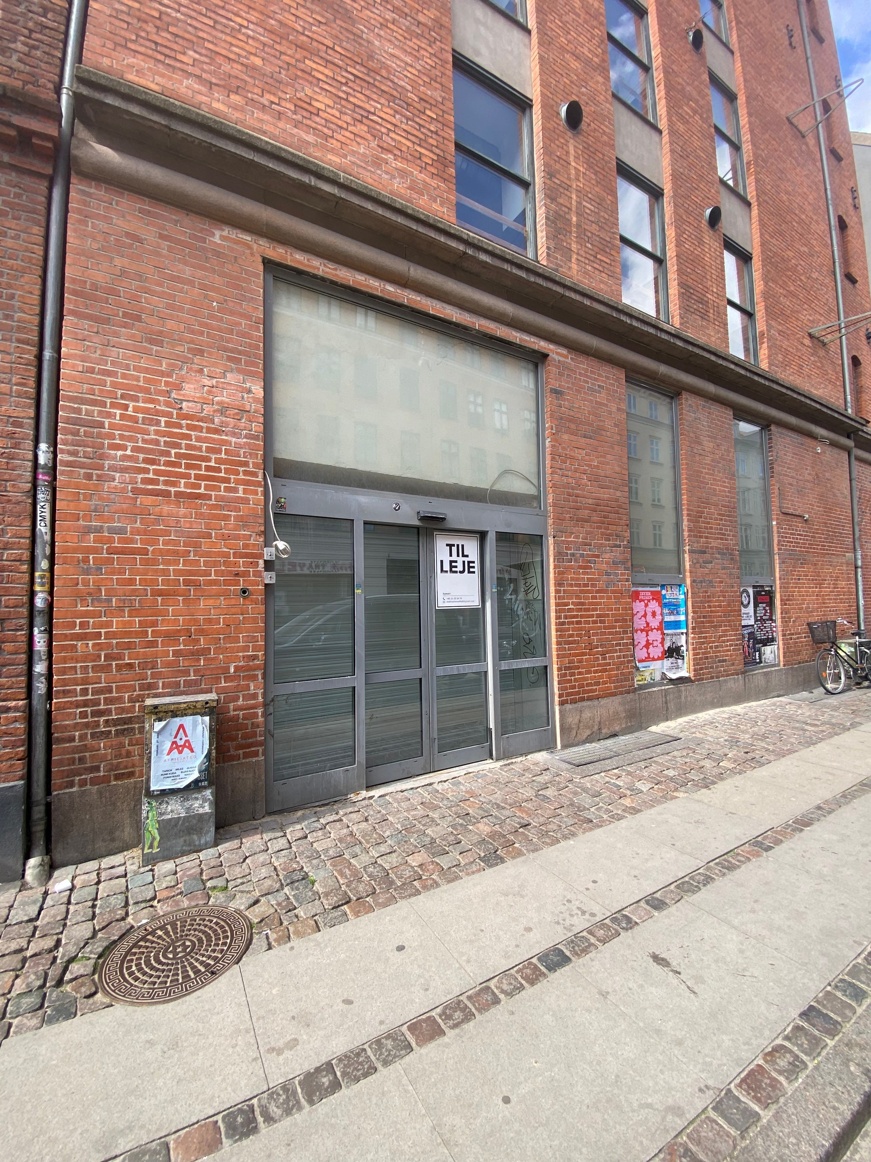 Rød teglstensbygning med indgangsdør, skilte og flere vinduer og reklameplakater på hovedgade