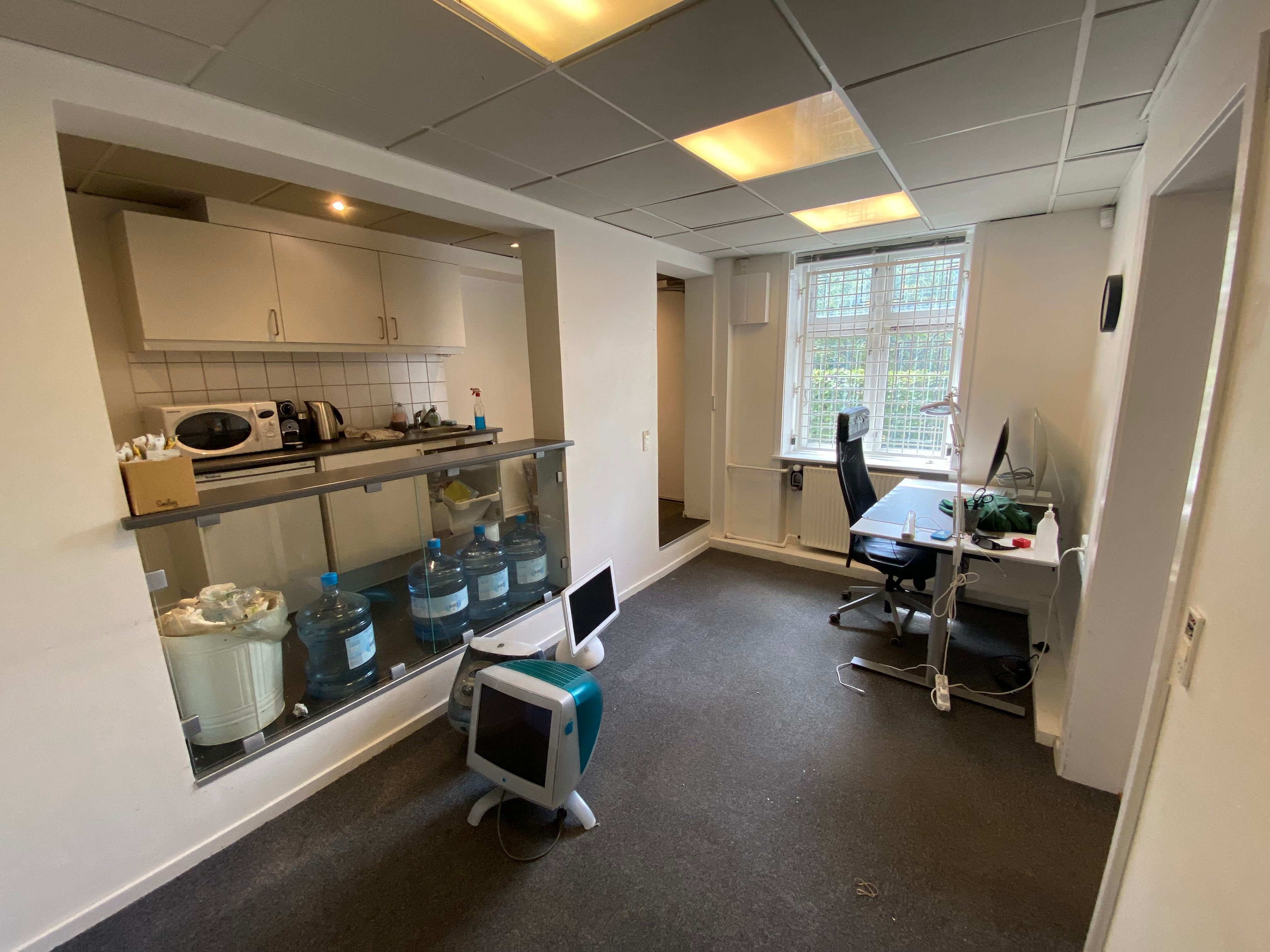 Et kontorlokal med to skriveborde, computere og en mikrobølgeovn på en hylde. Under hylden er der en papkasse og en kasse