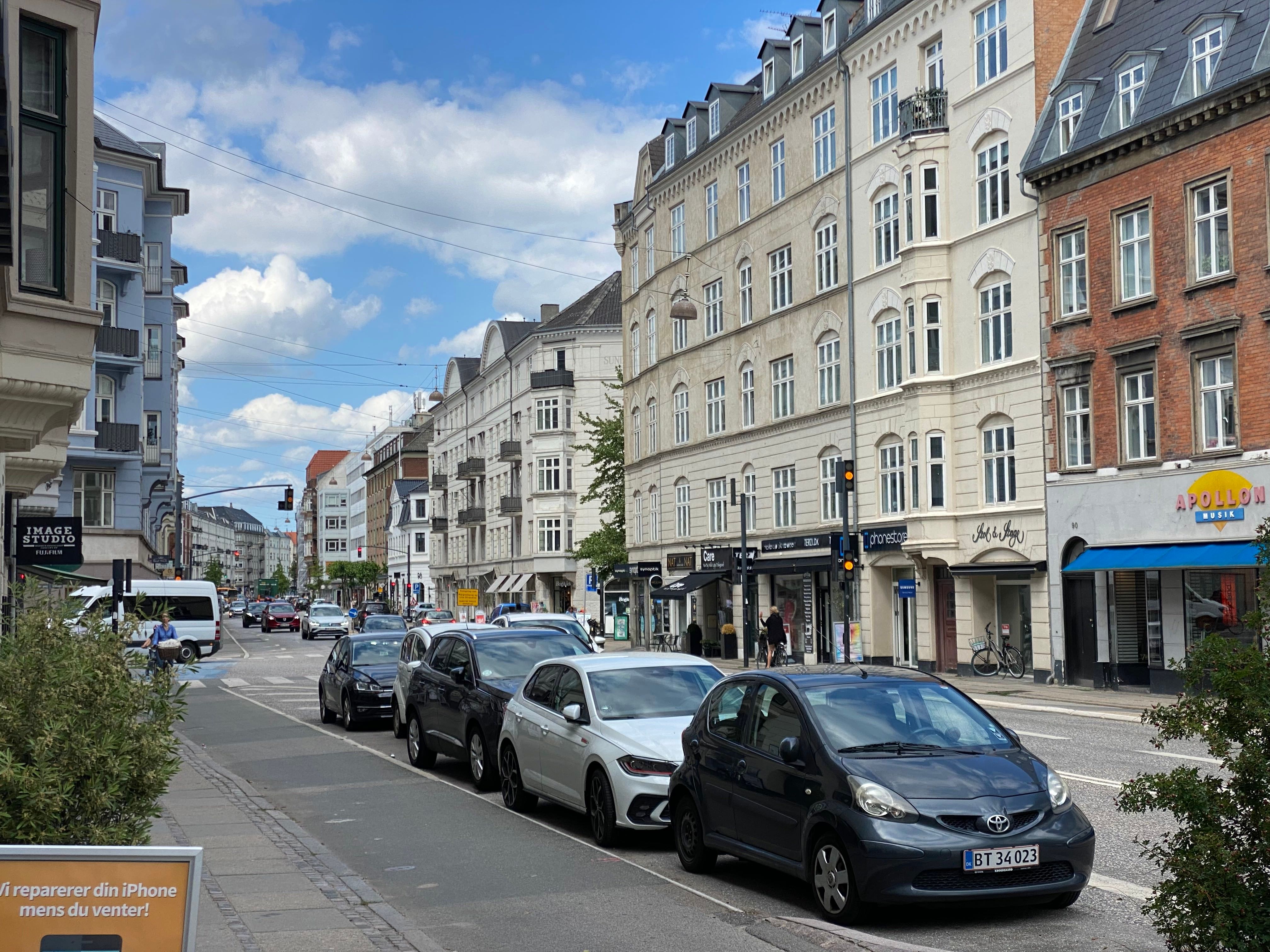 Parkerede biler på en gade i København, med folk og cykler langs fortov og forretninger med solbriller og højtalere