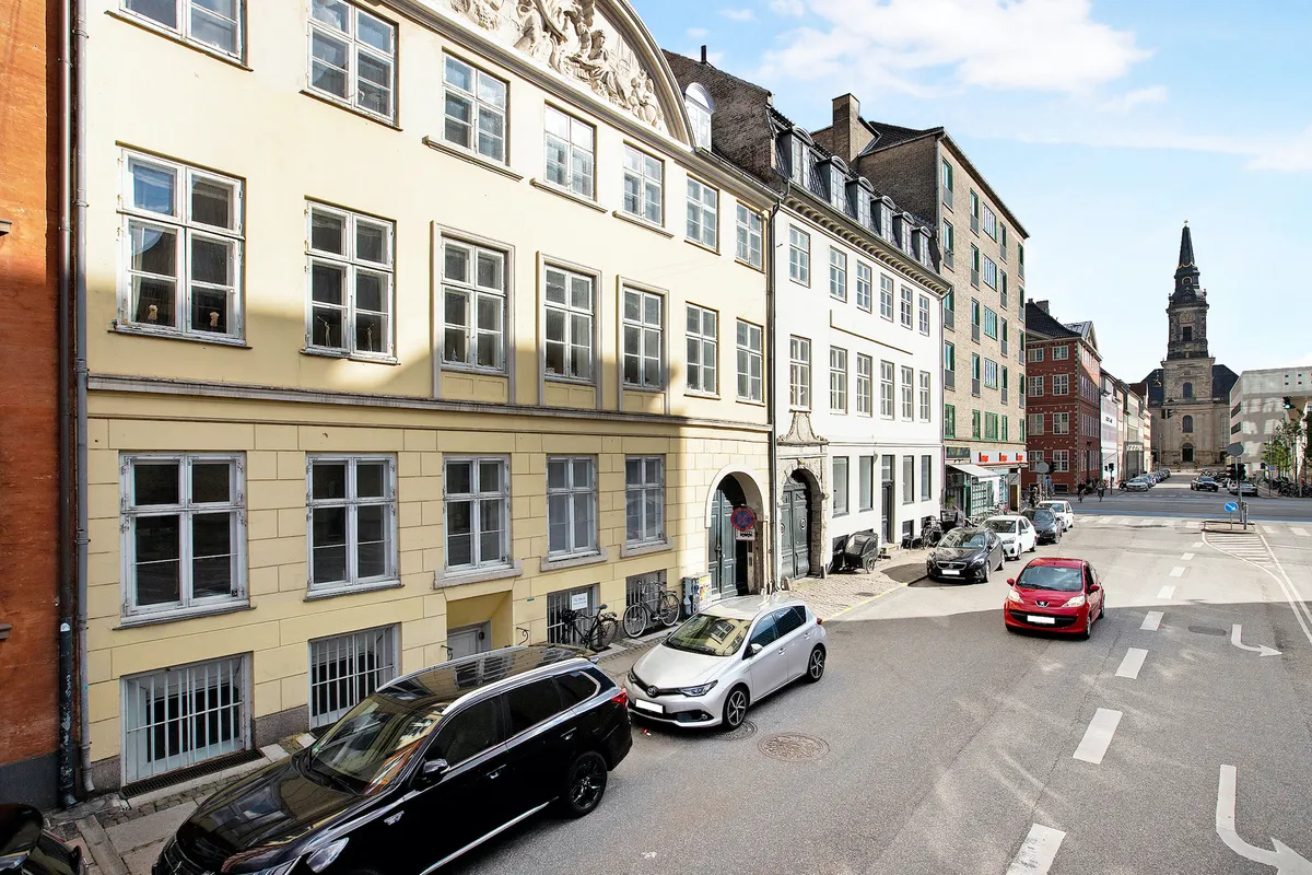 To biler parkeret i en gade med udsigt til en gammel bygning med flere vinduer og en kirke i baggrunden