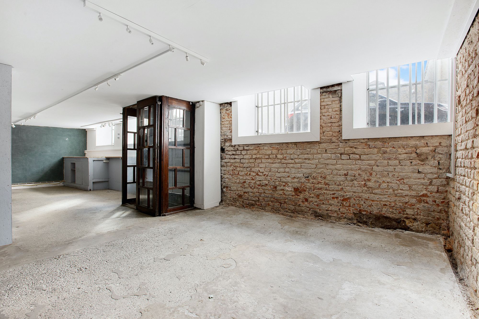 Stor tømmerkælder med højloft, to vindu og en dør. Væggen er af røde mursten og har to vinduer med stænger. Loftet er hvidt