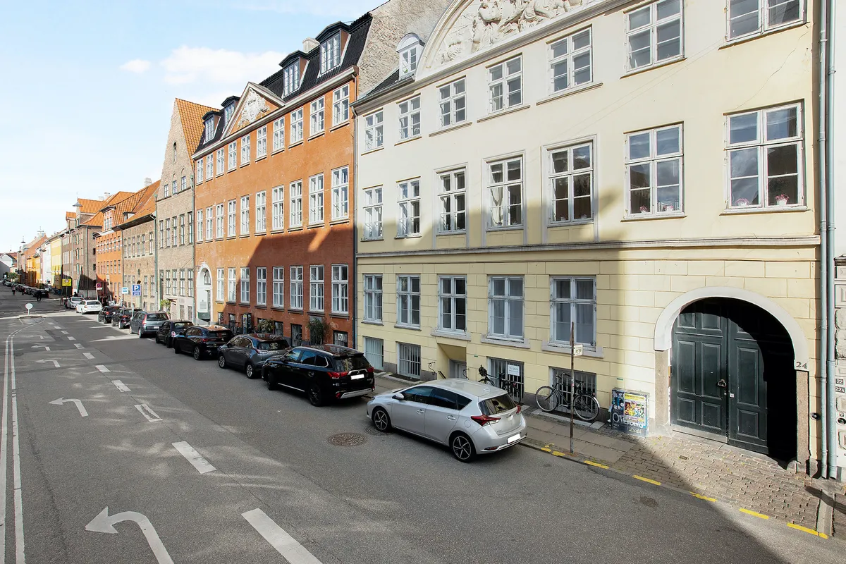 En lille gade i København med parkerede biler og en gågade med gamle bygninger med røde tag og store vinduer
