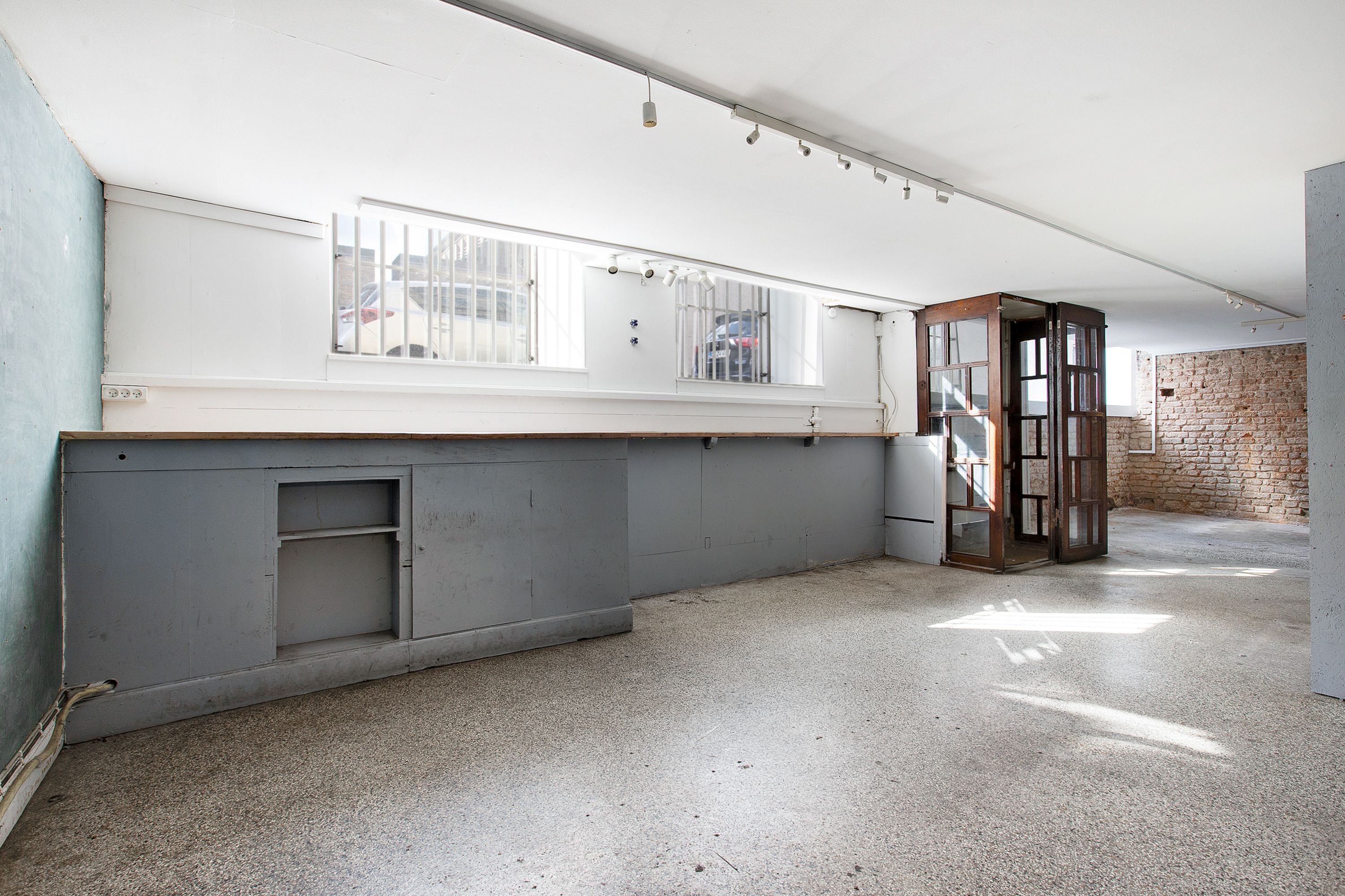 Lokal med lyse gulve og loft, samt to vinduer med stående jaluser. Lysslanger hænger på loftet, og der er en åben dør til et