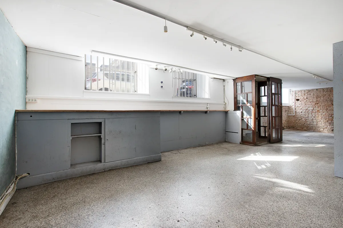 Lokal med lyse gulve og loft, samt to vinduer med stående jaluser. Lysslanger hænger på loftet, og der er en åben dør til et