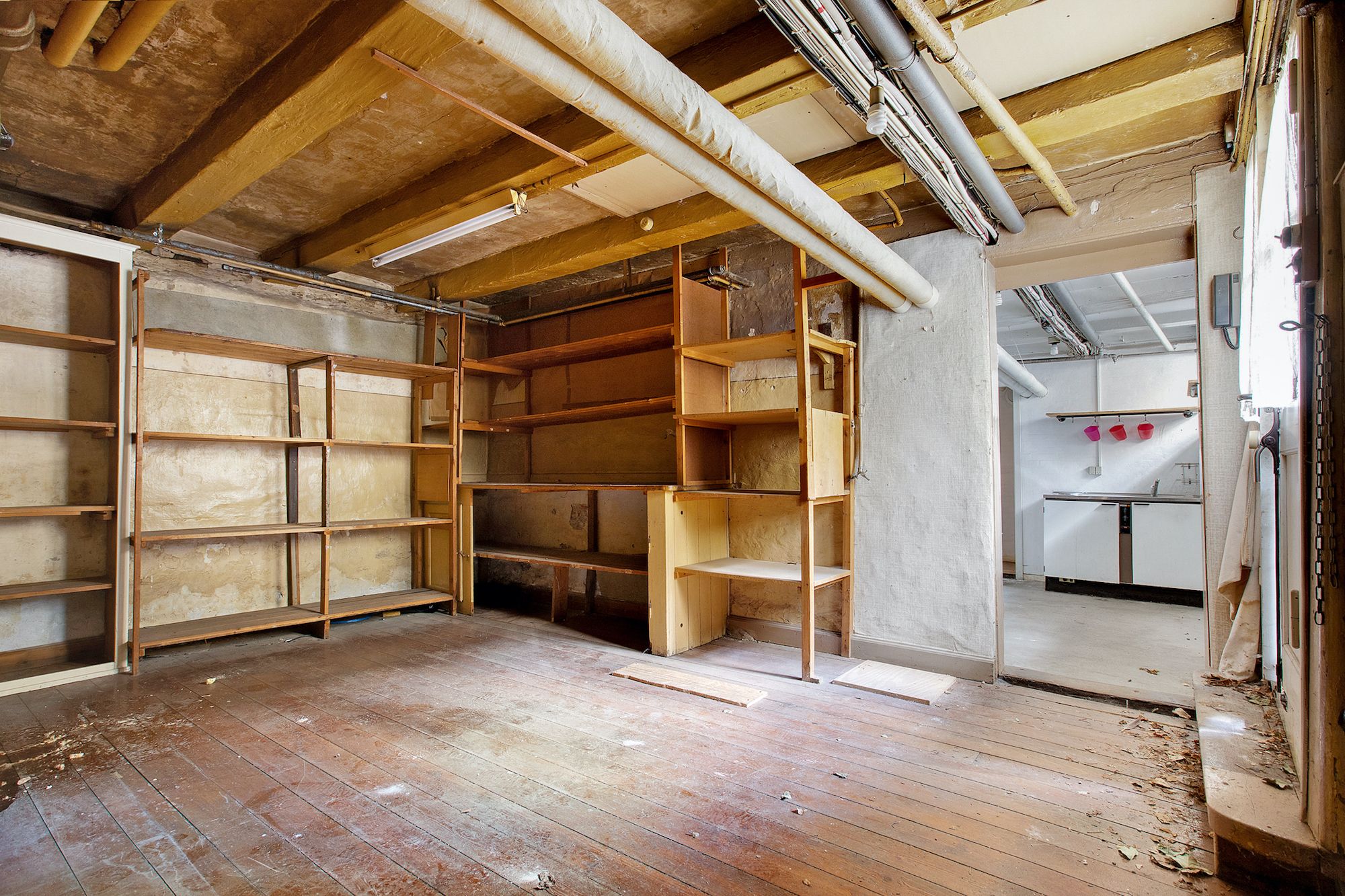 Forladt stue med trægulve og træhylder. Lampe og rør på loftet. Åbning til køkken med vaskebækken og rød opvask.