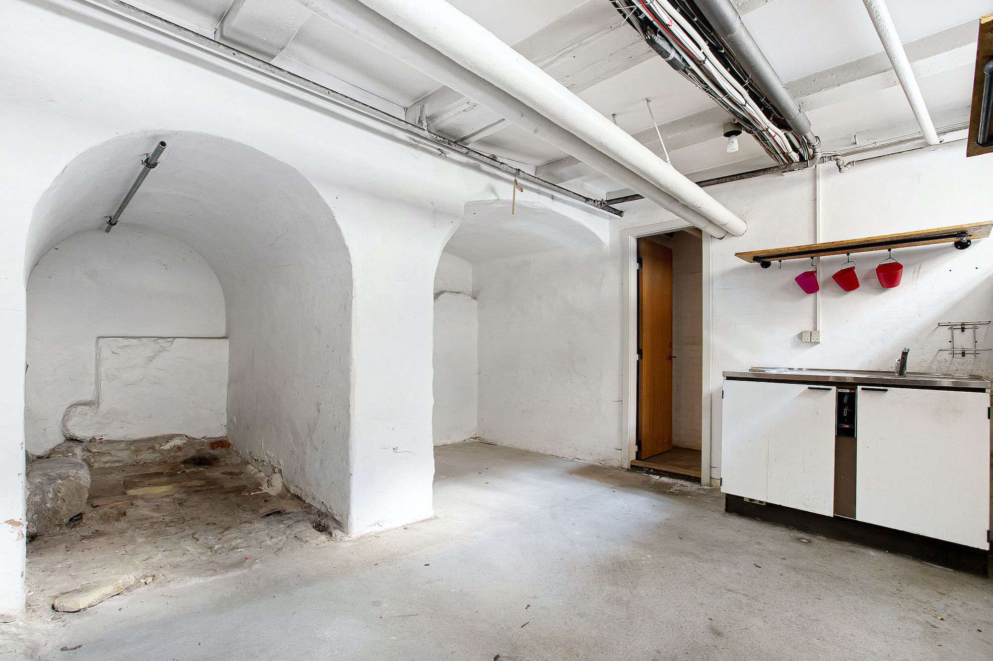 En gammel kælder med højt hvælvede stenvægge og lyse lofter samt rør og elektriske ledninger og nogle røde spande hængende