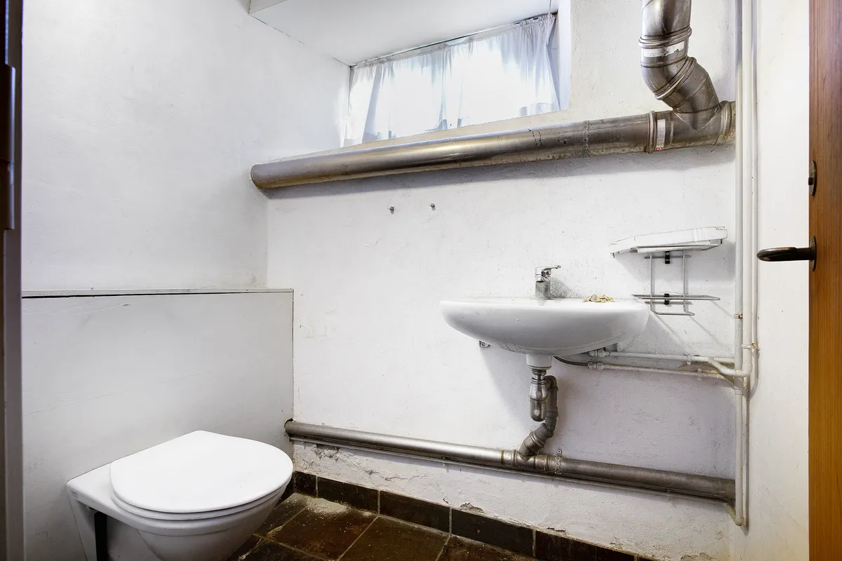 Toilet med forældet interiør og udsugningsrør i loftet og ved væggen samt et højt vindue med gardiner