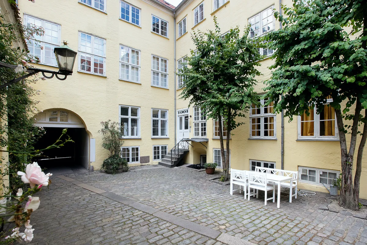 Lysgul bygning med flere vinduer og en åbningsportal, hvor der er en række hvide stole og et bord uden for