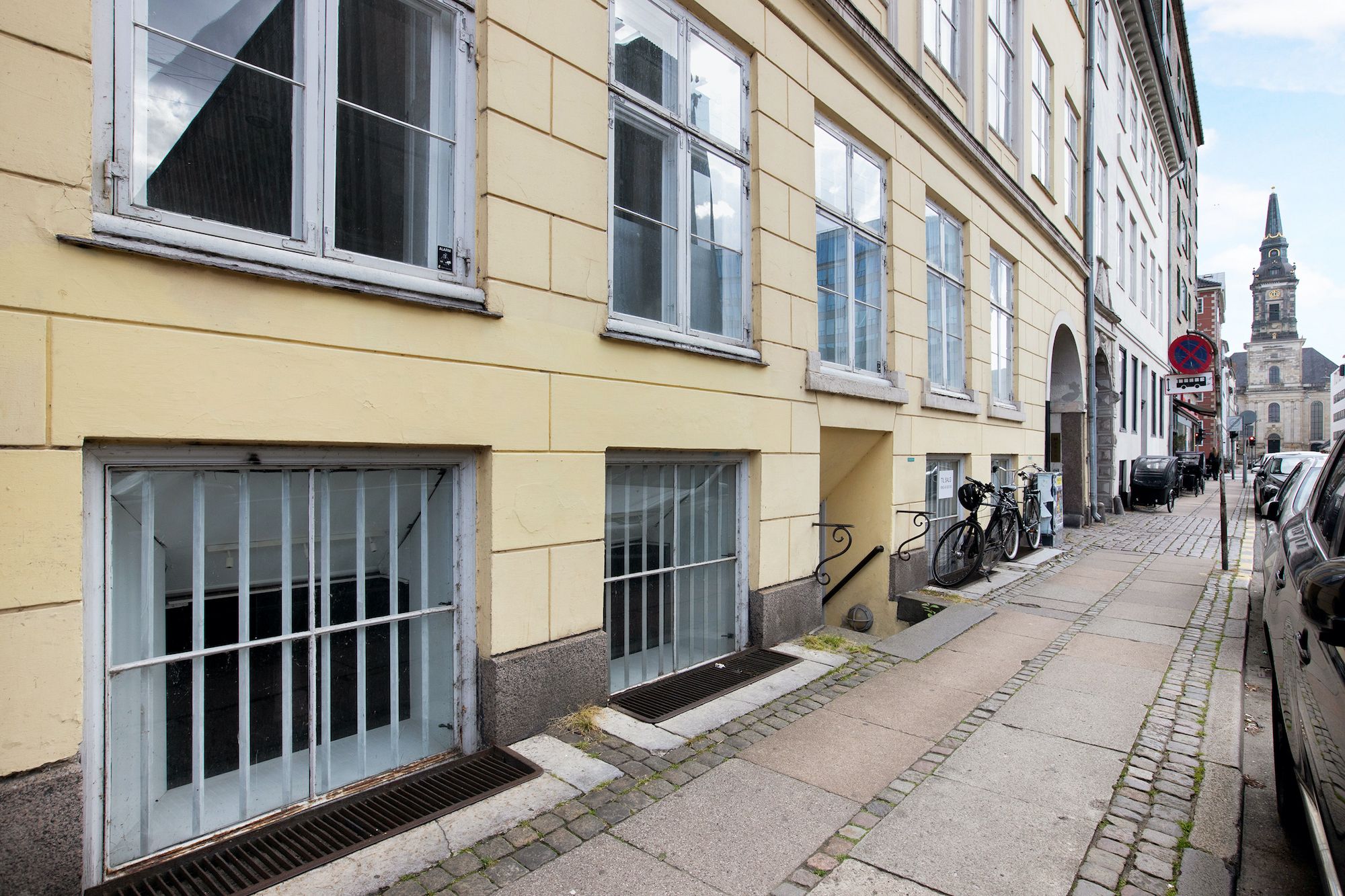 Vej med gul murstensbrolægning og parkerede biler, gamle bygninger med flere vinduer og glasdøre, udsigt til kirke og rødt