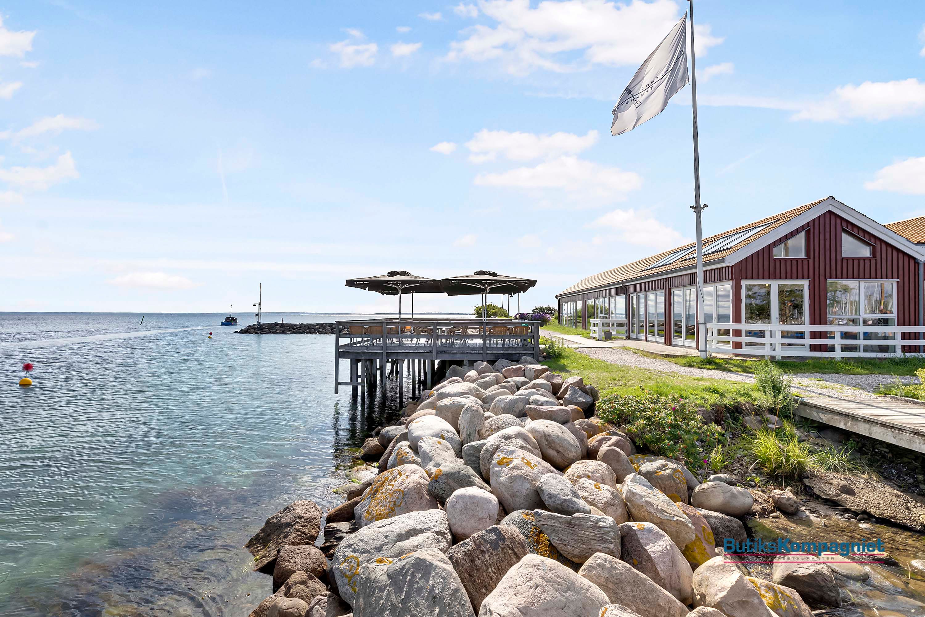 Langs stranden er der en havudsigtsrestaurant med flag, trægulve og grønne haver med blomster.