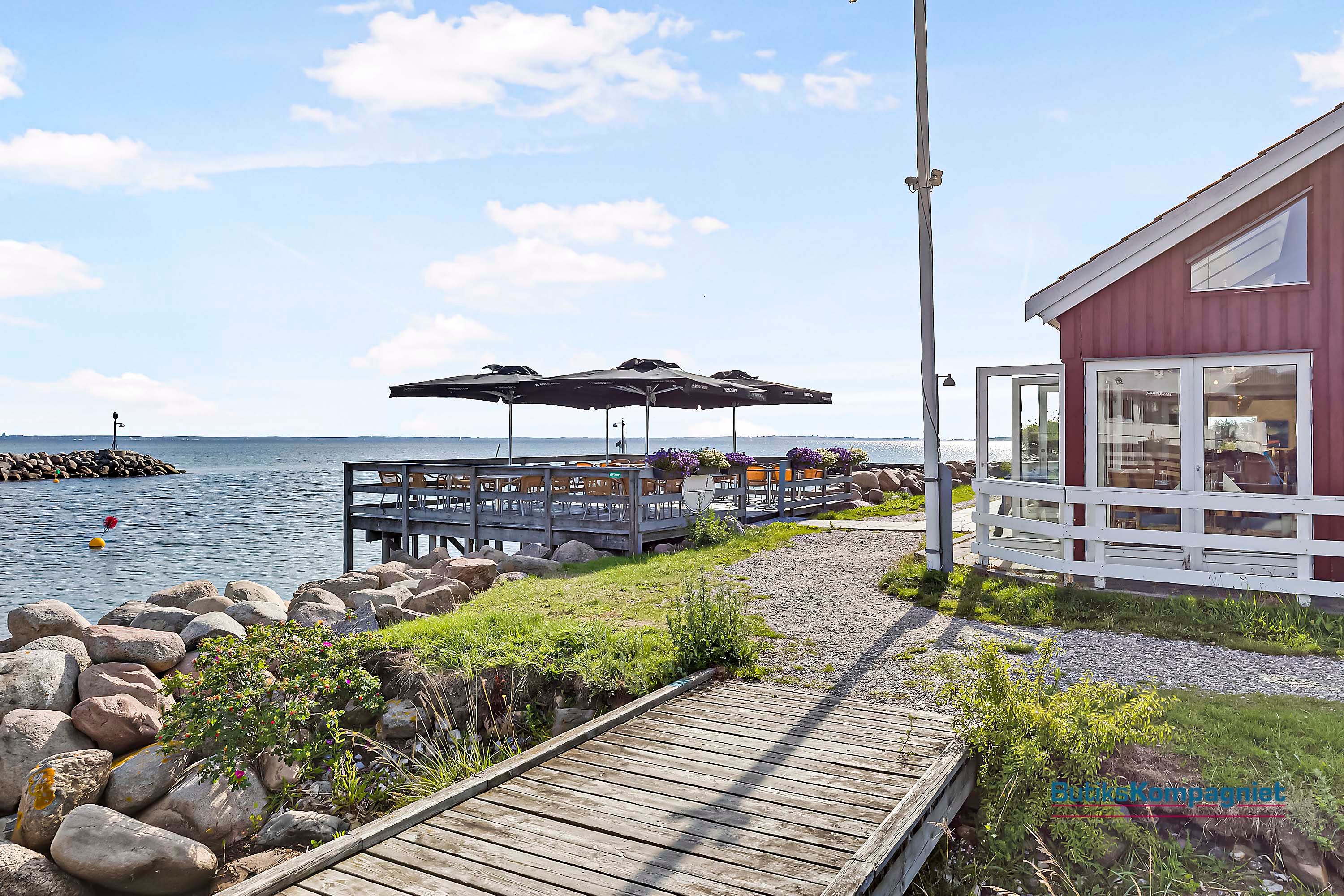 En række sædestole under parasoller og planter på en træbro med udsigt over Nivås havn.