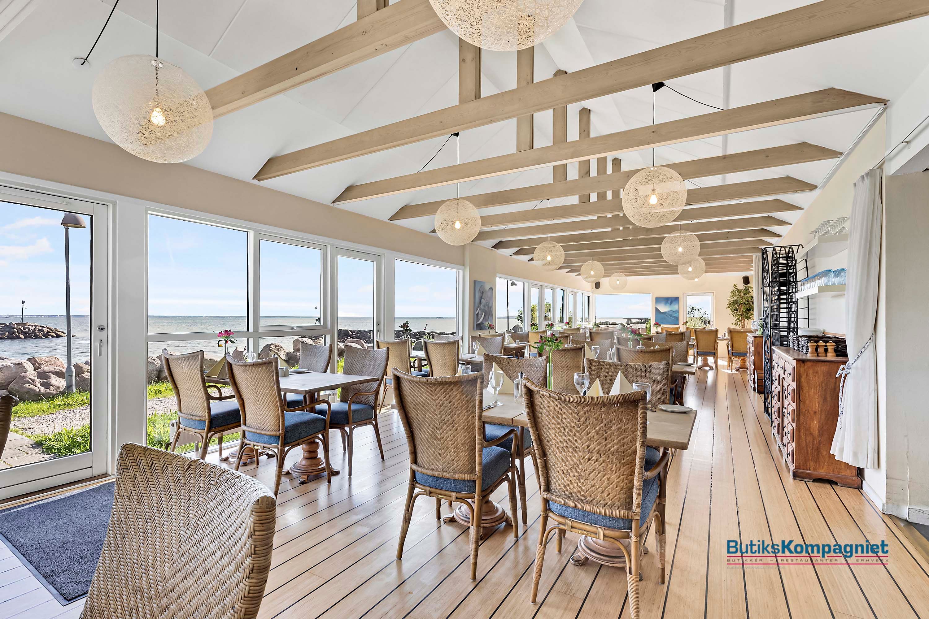 Elegant restaurant med havudsigt, lyse loftsbjælke, trægulve, store glasvinduer og træstole med blå pudsede sæder