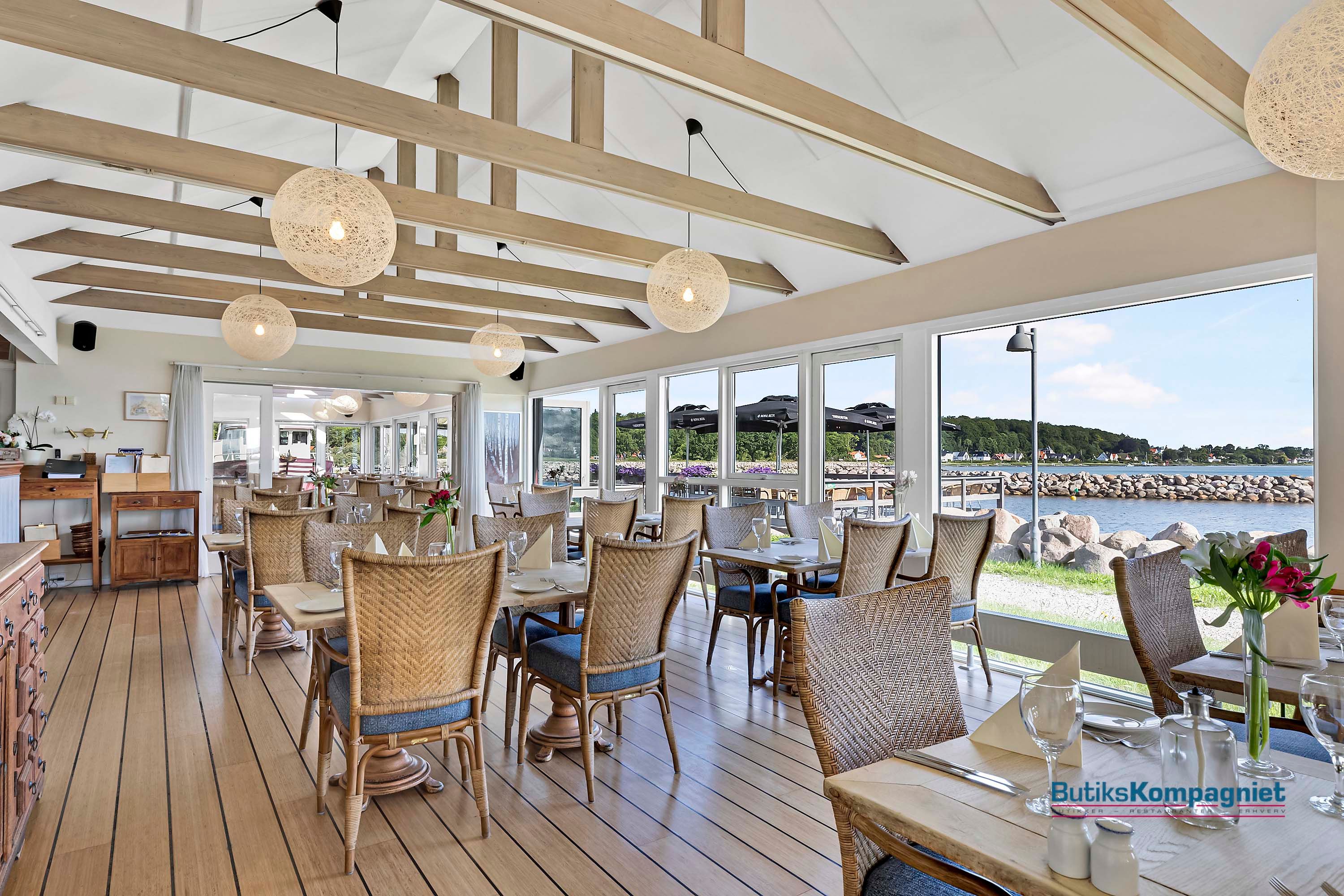 En restaurant med trægulve og havudsigt, hvor lysekroner hænger fra loftet, og søvogne står udenfor