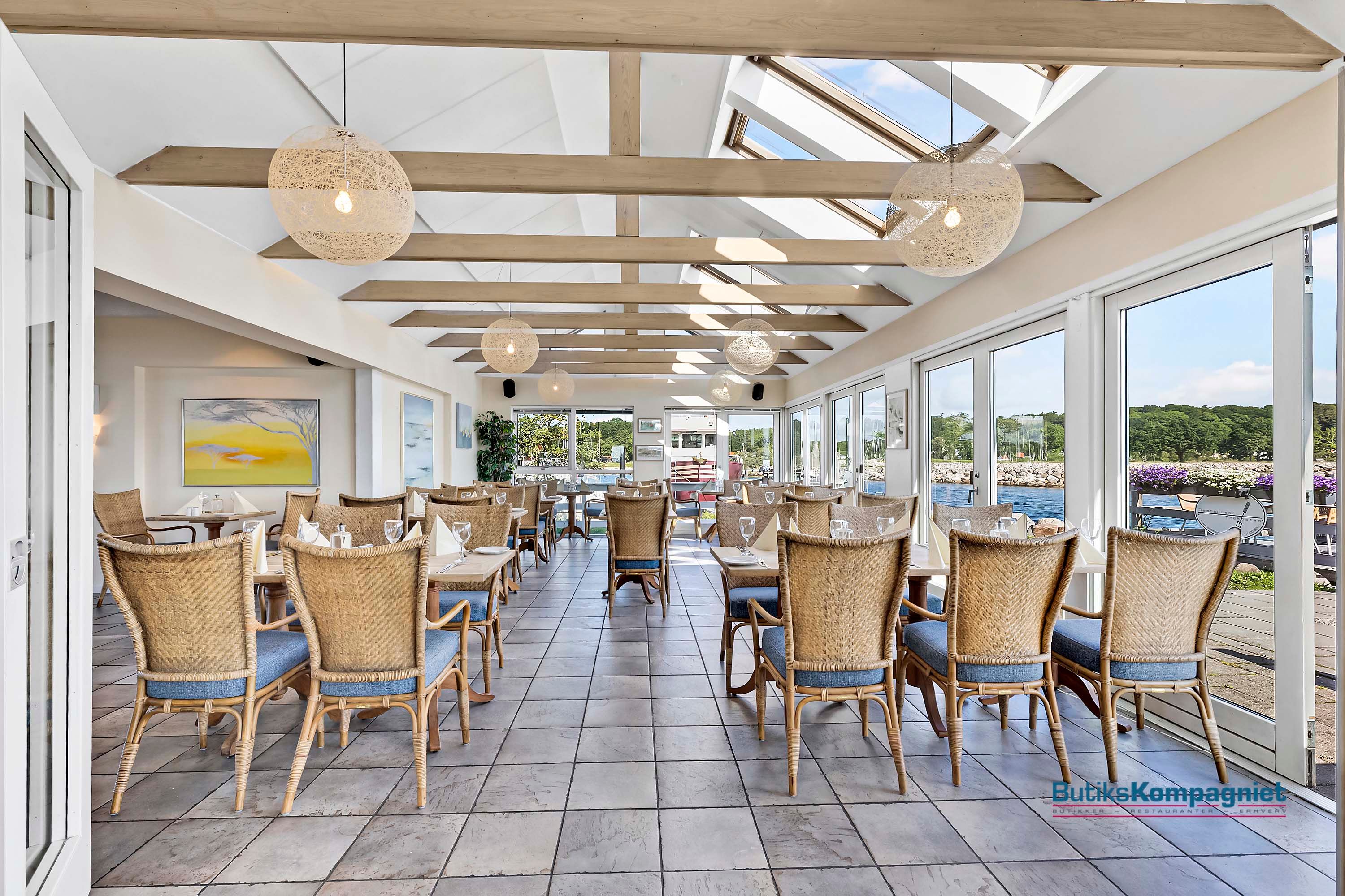 En restaurant med lyse loftsbjælker og vinduer med havudsigt og solrig himmel, smukke lysekroner og træstole med blå pudser