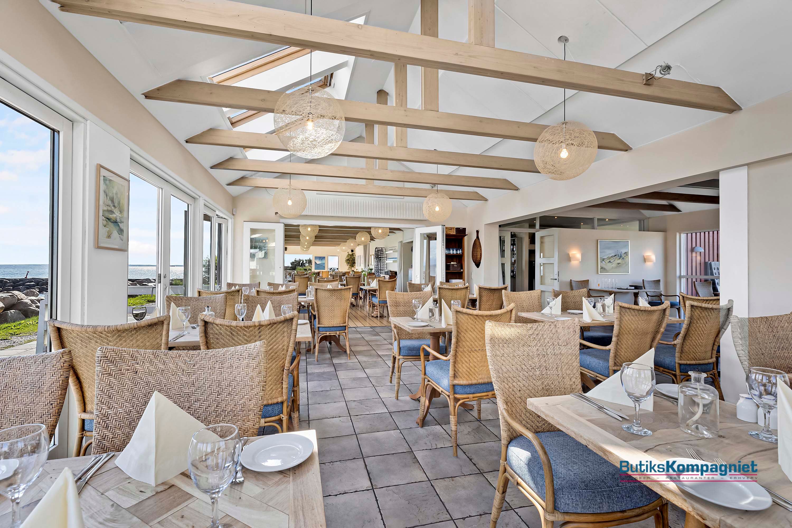 Indenfor moderne restaurant med havudsigt, hvor lyse bjælker stiger mod loftet, står flotte borde med vinglas og servietter