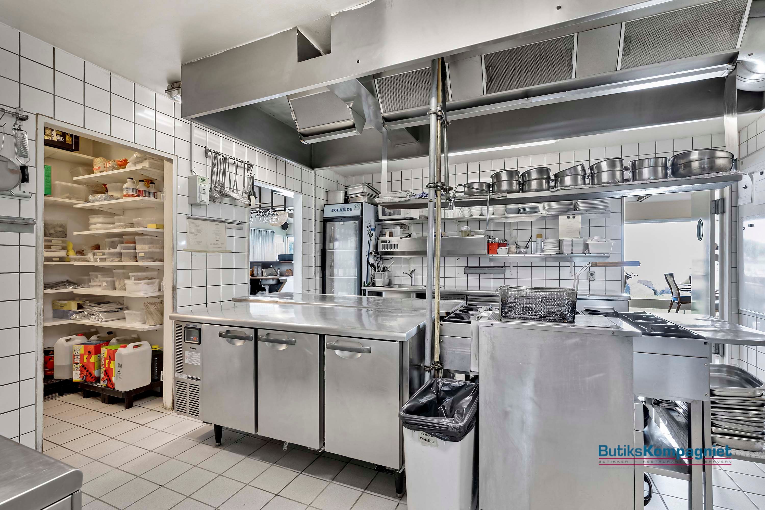 Professionel kommerciel køkkenudstyret med flere stålapparater, gryder, trækasser og skraldpose. Et lukket køleskab og en