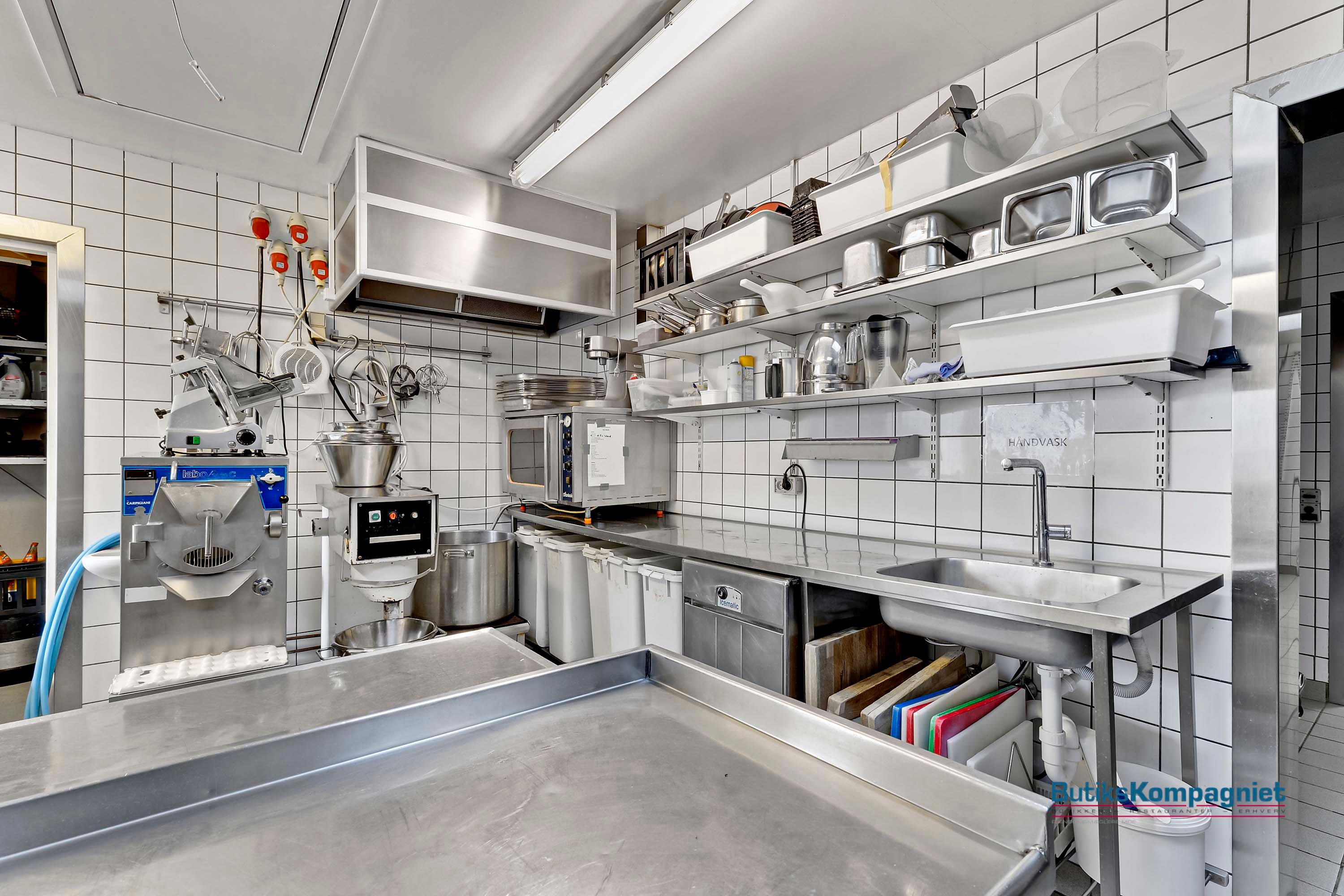 En stor kommerciel køkken med flere apparater og faciliteter, en stålbøjle og et blikloft. Der er en mikrobølgeovn og et