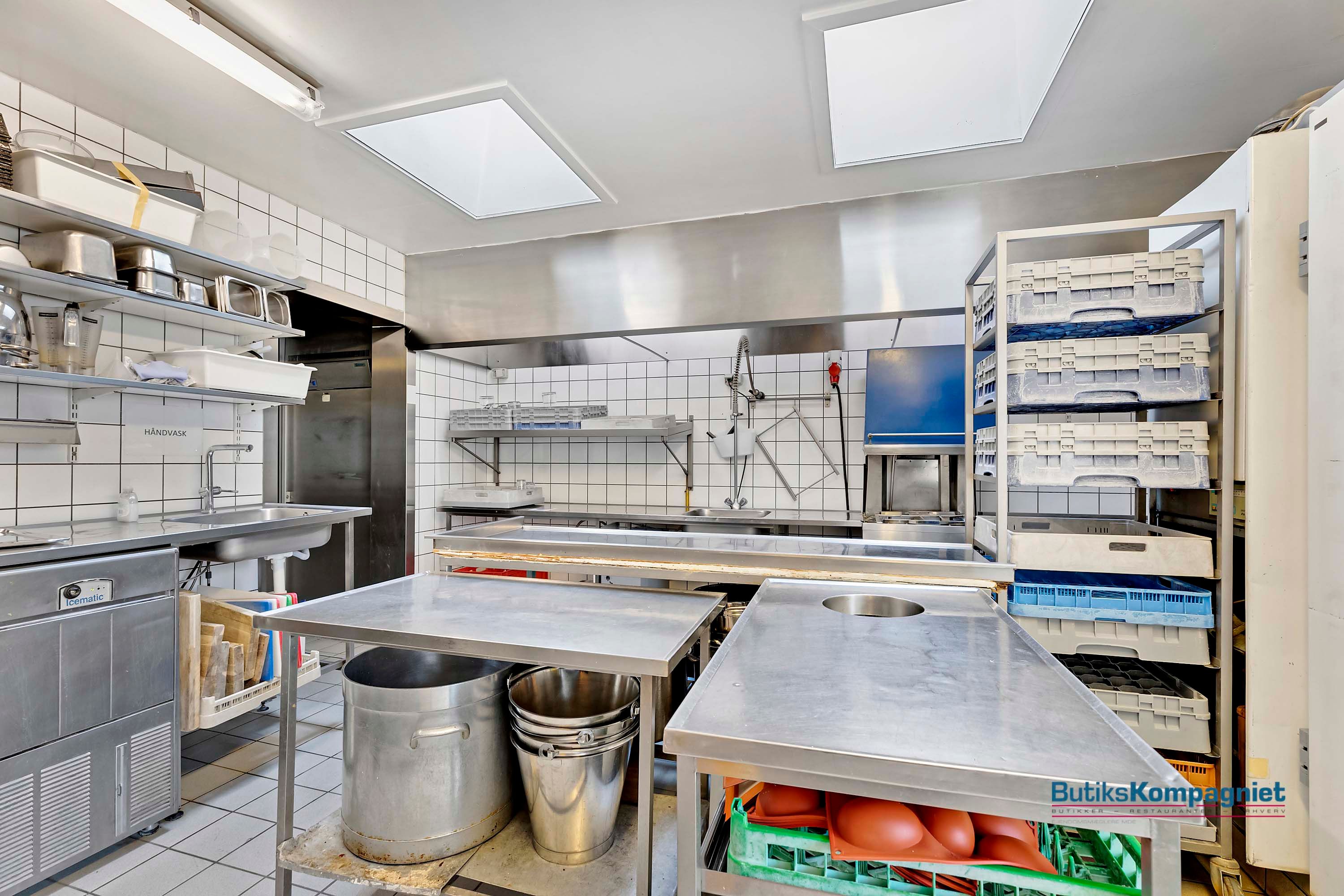 Moderne køkken med stålborde, metalbøjler, kassevogn og højloftsventilation i en professionel køkkenindretning. Et stort