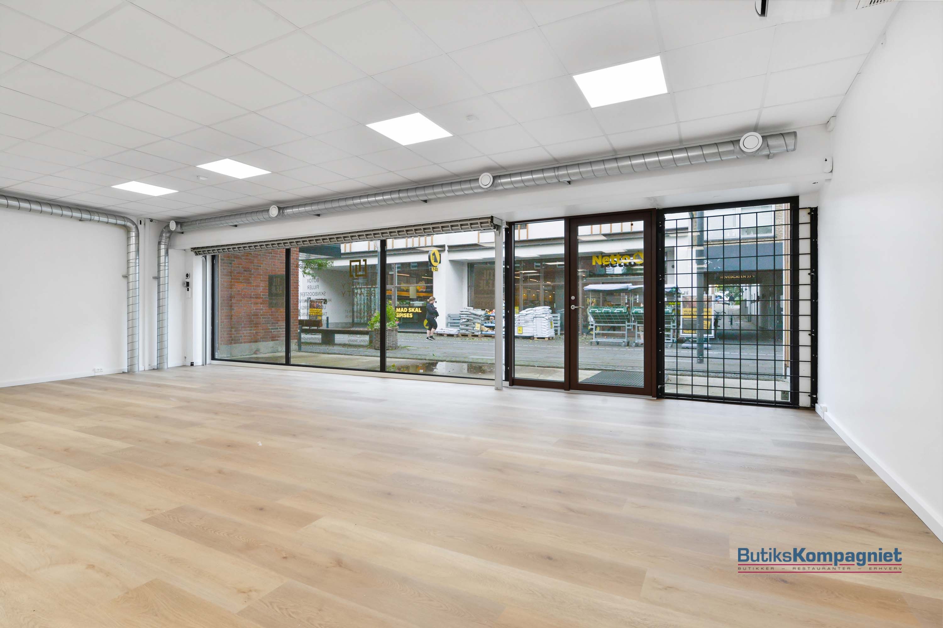 Butikslokaler med store glasruder og trægulv, der vender ud mod en gade med biler og folk.