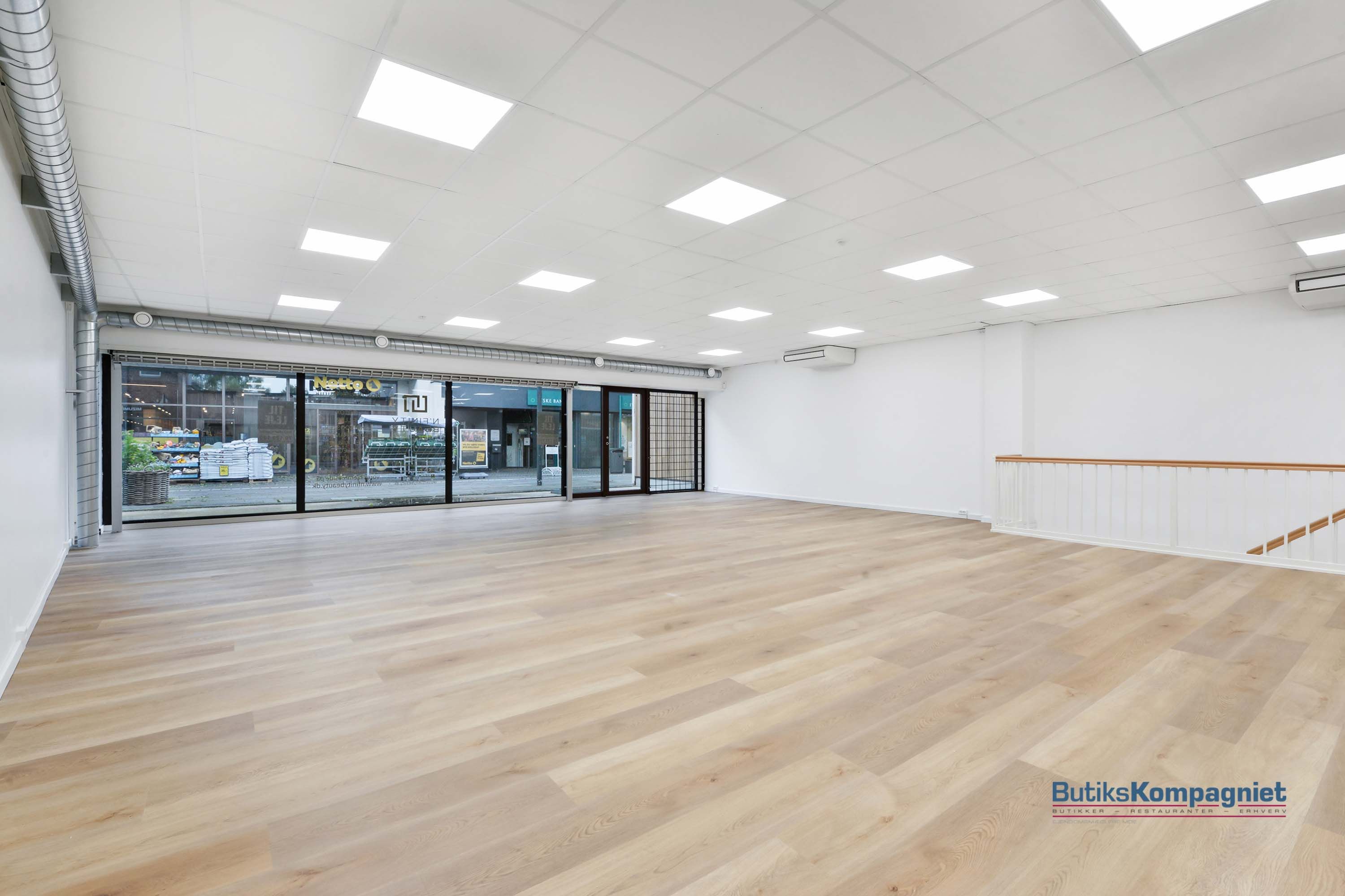 Ledig forretningslokale med store ruder og højloft, hvidt loft med flere indbyggede lys, lysegul trægulv og hvid væg, og
