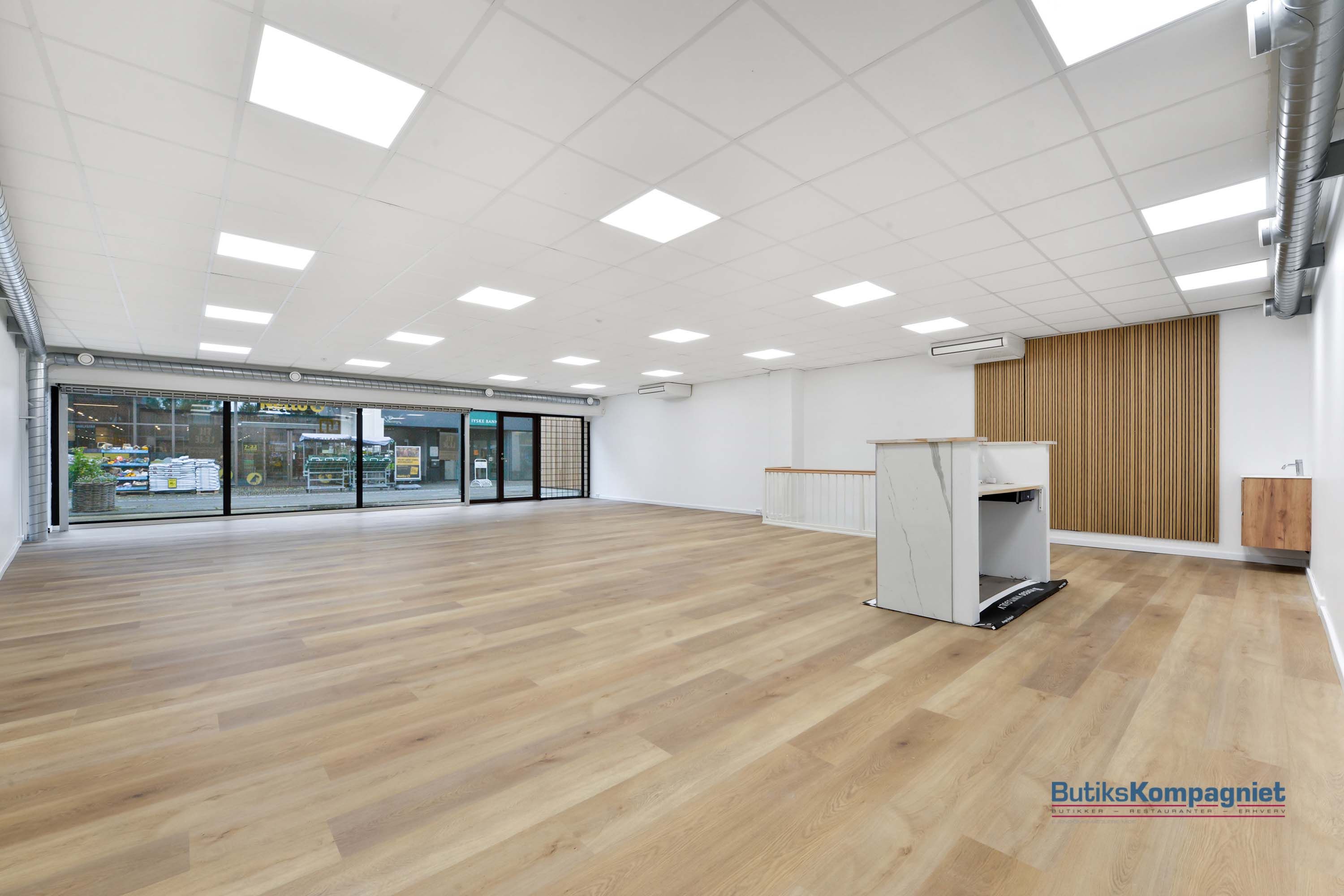 Store tomt butikslokaler med lyse loftspaneler, hvide vægge, træbeklædte vægge, marmordisk og lyse trægulve.
