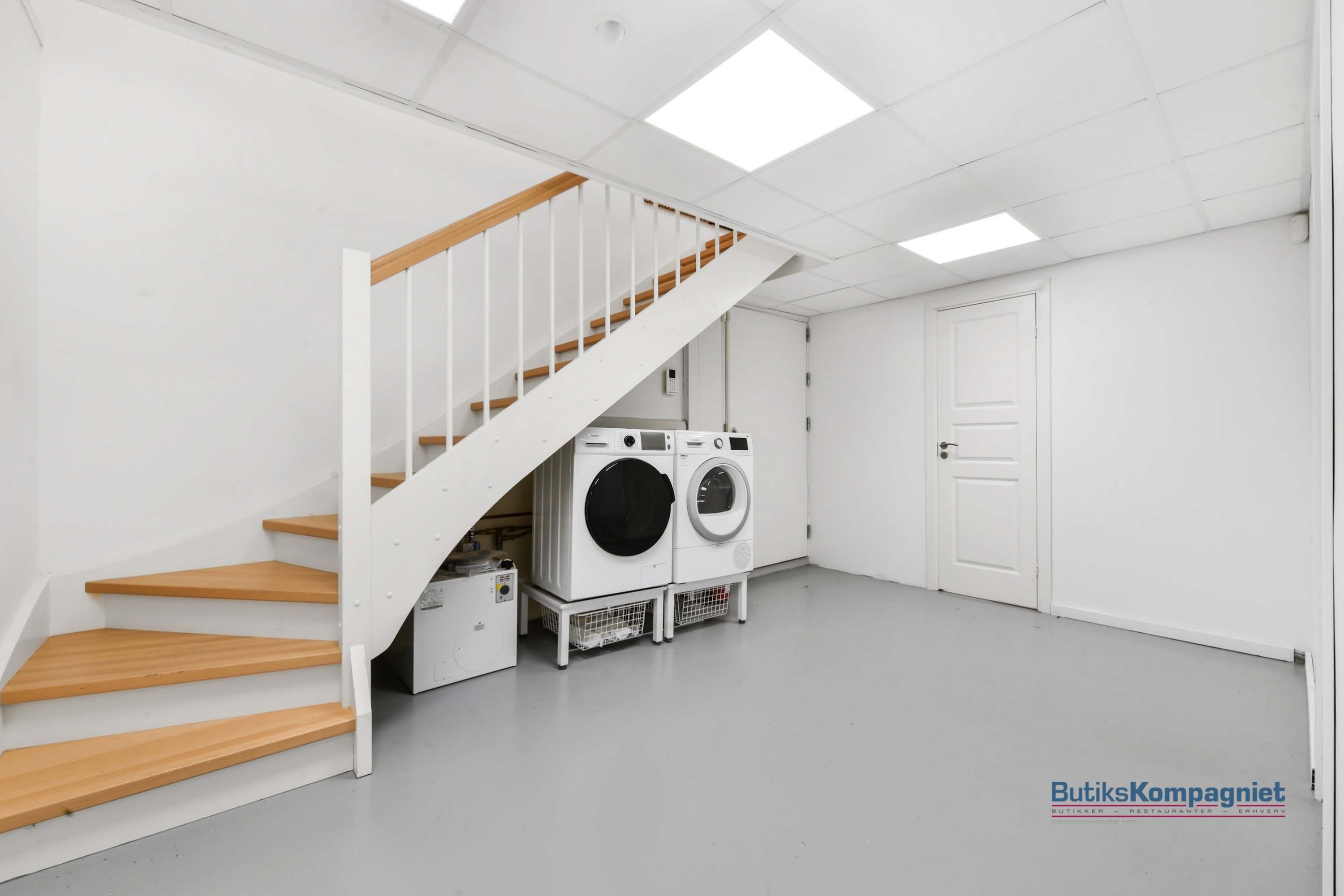 En rummelig kælder med hvide vægge, gulv og loft samt en hvid trappe og to hvide vaskemaskiner
