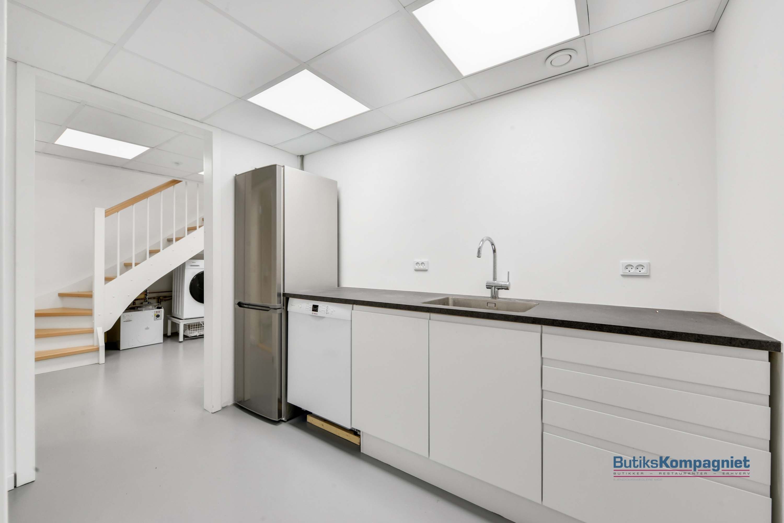 En moderne køkken med lyse vægge og højloft, integreret vaskemaskine og tørretumbler, og en stor køleskab samt en strygebord.