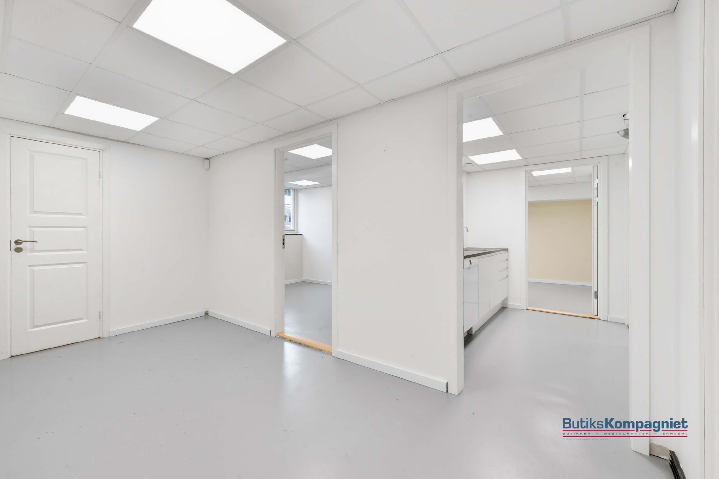 Et rent og ubenyttet kontor med lyse loftslampe, hvide vægge og gulve samt to døre og et kammer