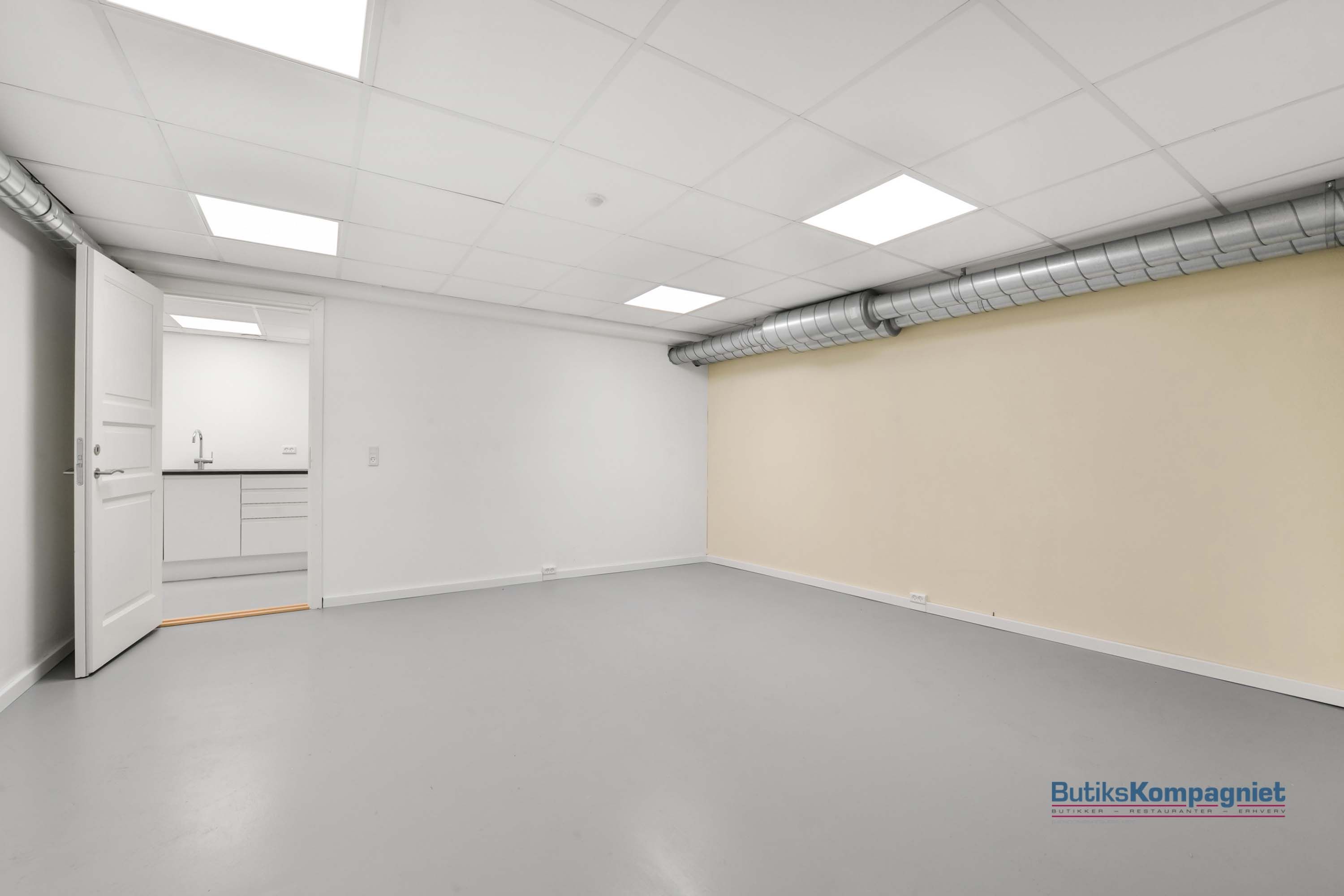 En tom, hvid og ren stue med højt loft og hvide vægge, samt en lysegrå gulv. Der er en åben dør, som fører til et køkken med